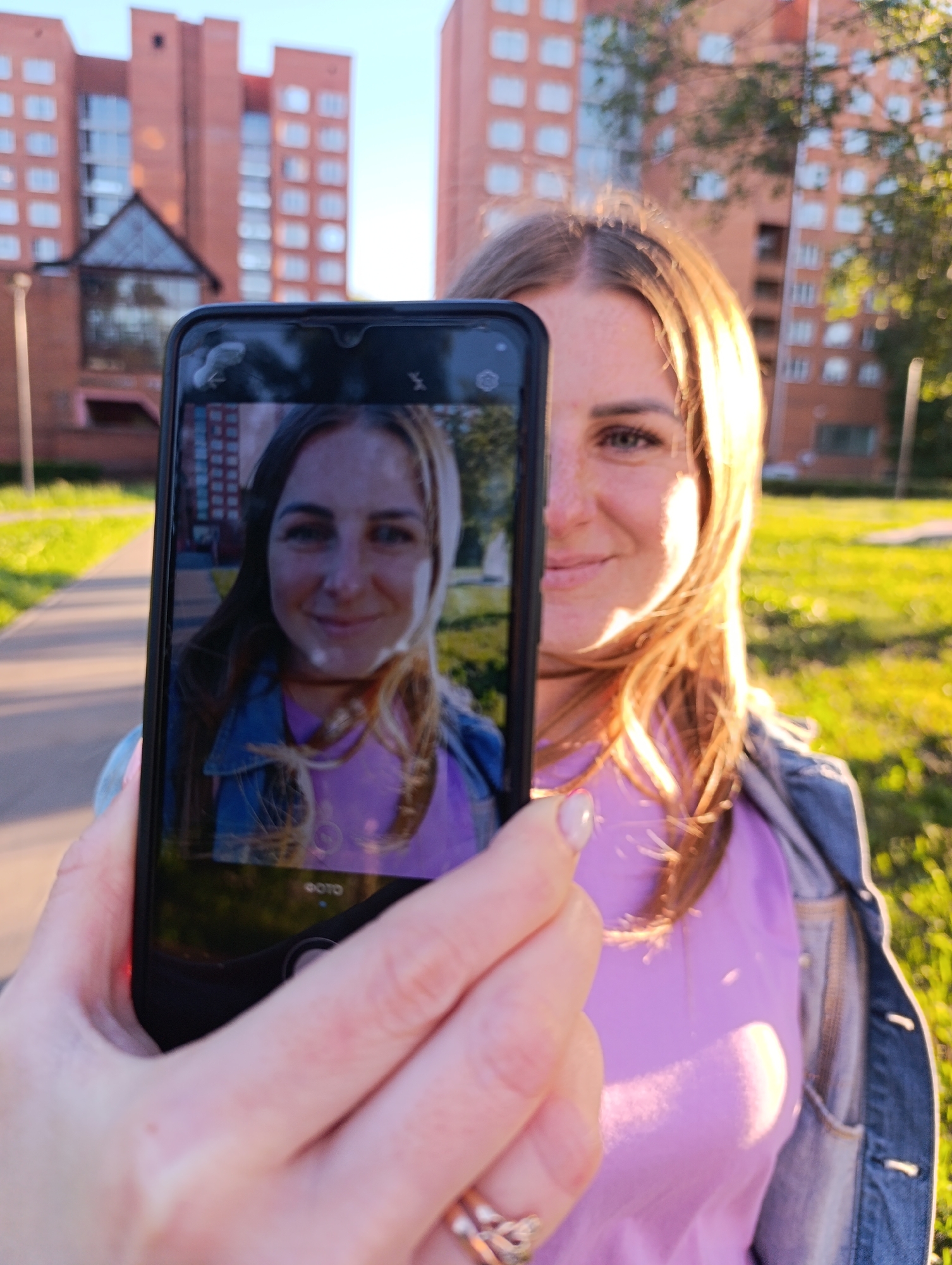 A young woman is taking a selfie with a smartphone. The phone is held up, obscuring the right side of her face. The phone screen displays a reflection of her face, smiling, with her hair down. She is wearing a pink t-shirt and a denim jacket. The background shows a brick building, possibly an apartment complex, with some trees and grass. The lighting suggests it is late afternoon or early evening, with the sun casting a warm glow on the scene. The location is identified as Chernogolovka, Russia. The woman has freckles and appears happy. There is some visible text on the phone screen, which appears to be Russian and says "ФОТО" meaning "PHOTO".