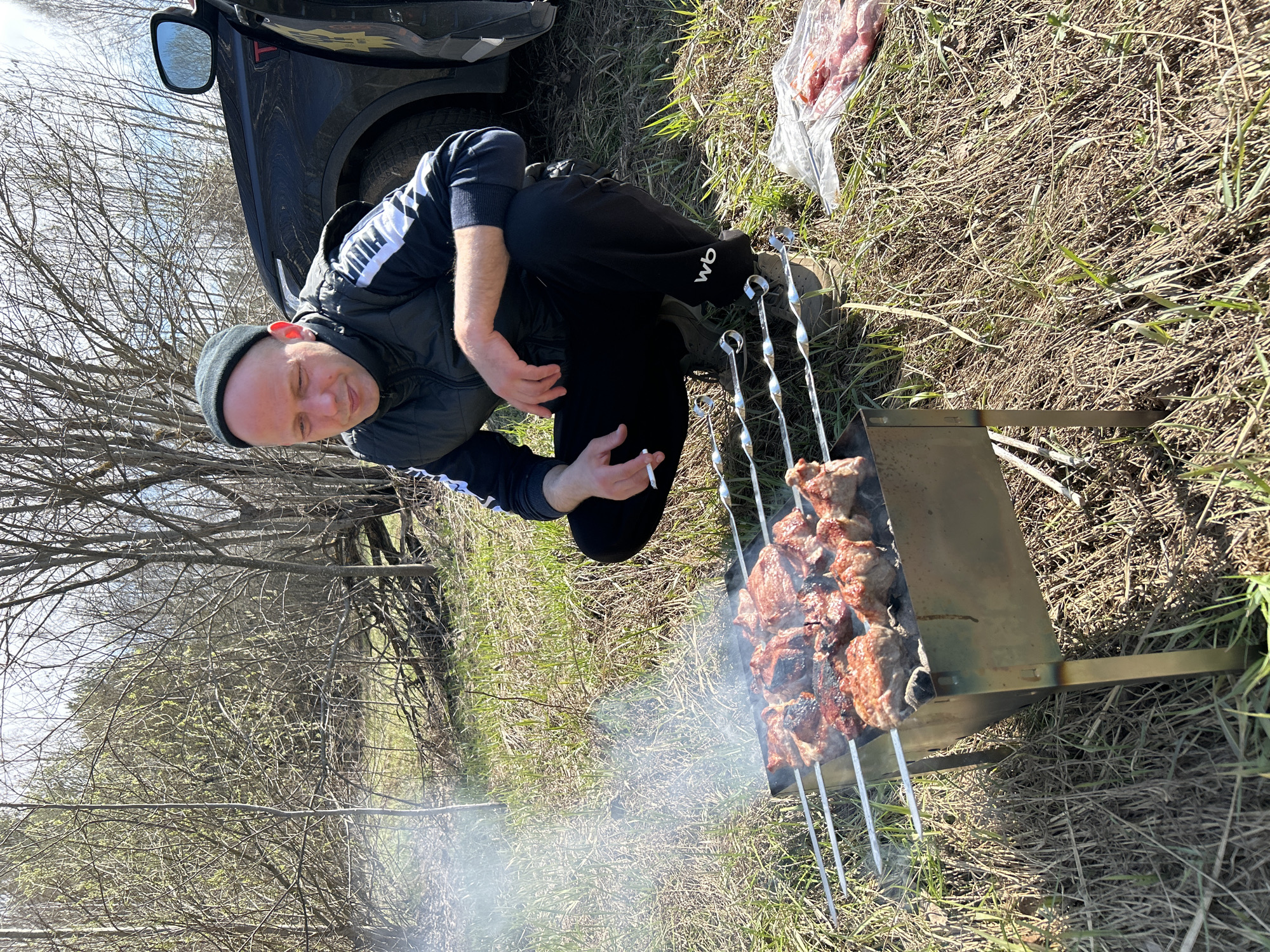 A photograph, oriented vertically, captures a man cooking kebabs outdoors. He kneels beside a barbecue grill on which several metal skewers hold pieces of meat. The meat closest to the viewer appears slightly charred. Smoke rises from the grill. The man holds a cigarette in his hand. He is wearing a dark beanie, a black jacket with white stripes on the sleeves, and black pants. Next to him, on the ground, is a plastic bag containing raw meat. A black car is partially visible in the upper left corner of the frame. The background features bare trees and dry grass. The setting appears to be an outdoor, possibly rural, location.