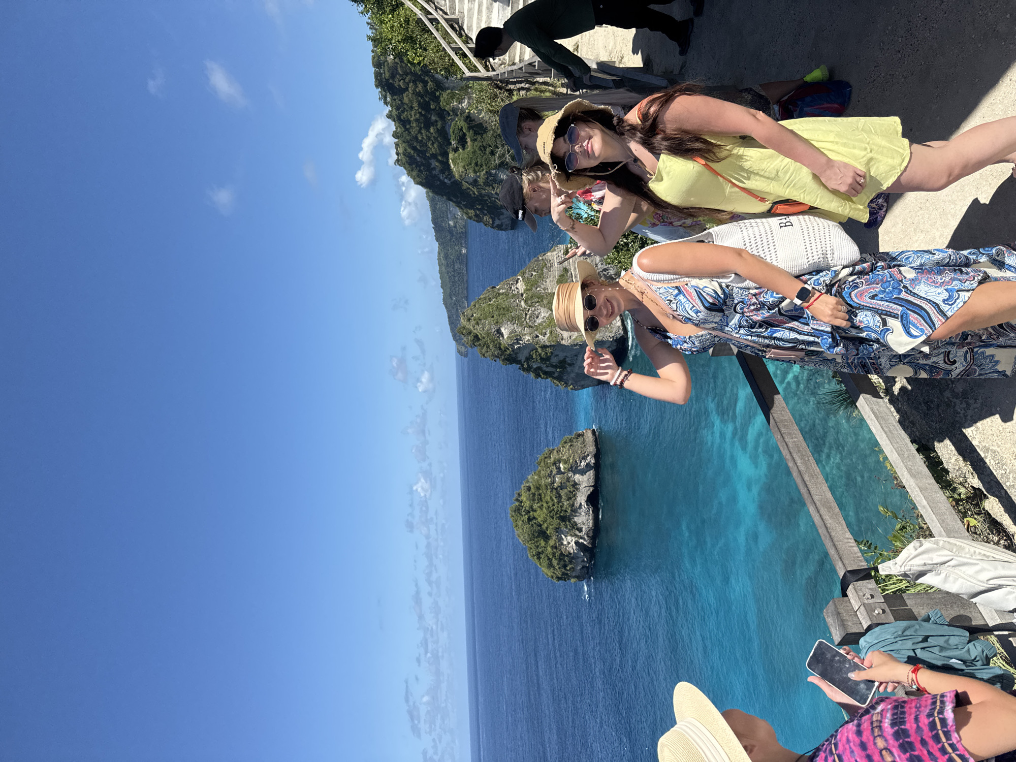 The image shows a group of four women posing for photos in Nusa Penida, Indonesia. The women are standing on a wooden platform overlooking a cliff and the ocean. The woman in the front is wearing a blue and white dress and a straw hat. The woman behind her is wearing a yellow dress and sunglasses. The other two women are standing further back, near a set of stairs. The ocean is turquoise blue and there is a small island in the distance. The sky is blue and sunny.
