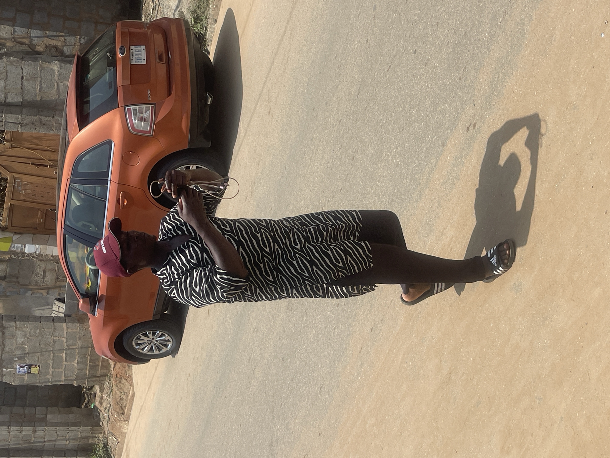 A person is standing on a paved road in what appears to be a neighborhood in Abuja, Nigeria. The person is wearing a black and white patterned tunic and dark leggings, with Adidas sandals on their feet. They are holding a coiled cable in their hands. Behind the person, an orange SUV is parked on the side of the road. The environment is sunny, with some buildings and a wooden gate visible in the background. The road surface is a mix of asphalt and dirt.