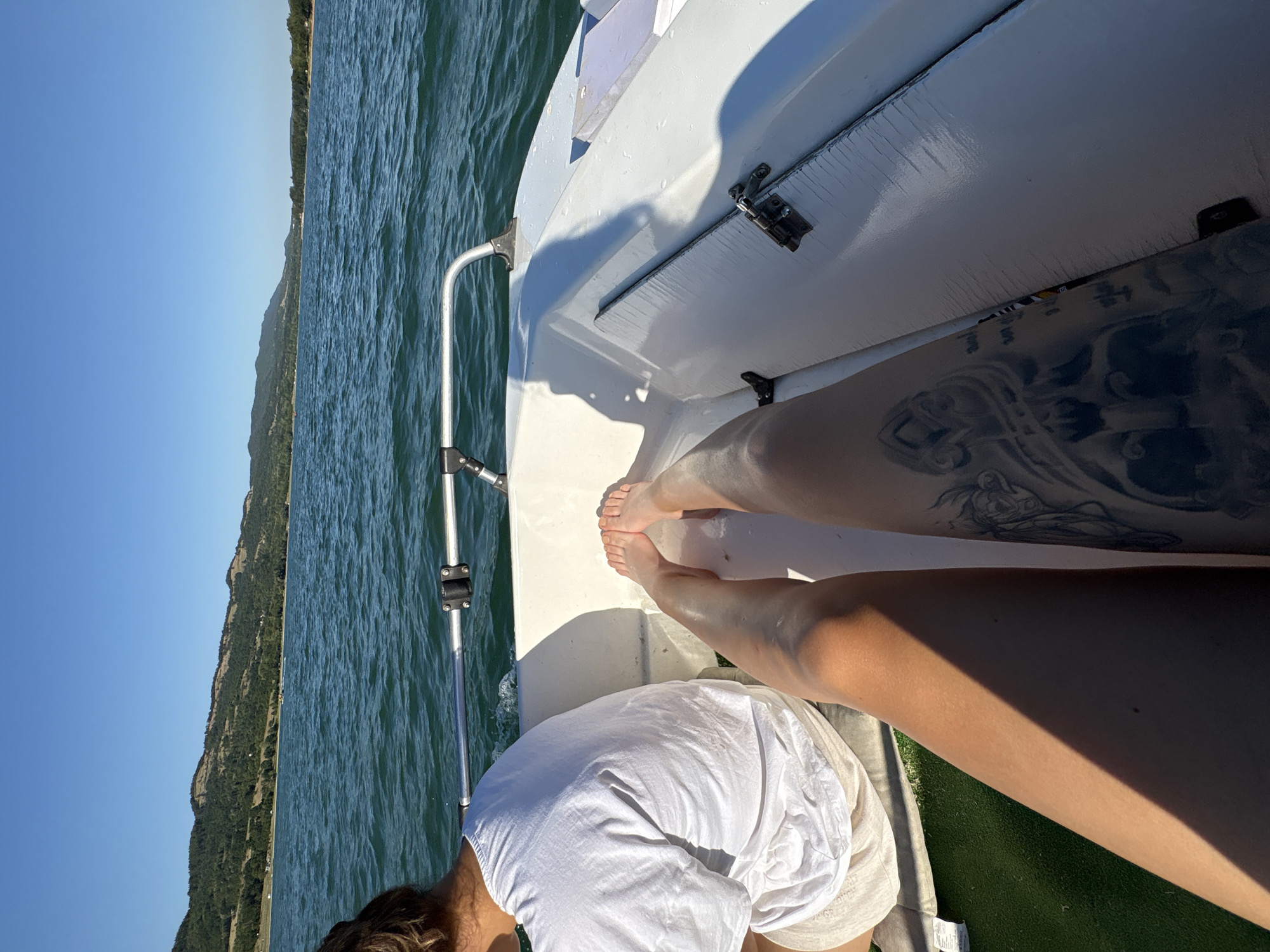 The image shows two individuals on a boat traveling on a body of water. One individual, seen from the back, wears a white t-shirt and light-colored shorts. The other individual's legs are prominently featured in the foreground, with bare feet and a tattoo visible on the left thigh. The boat appears to be moving, as evidenced by the wake in the water. The background consists of a hilly, tree-covered landscape under a clear blue sky. The location is identified as Golyama Zhelyazna, Bulgaria.