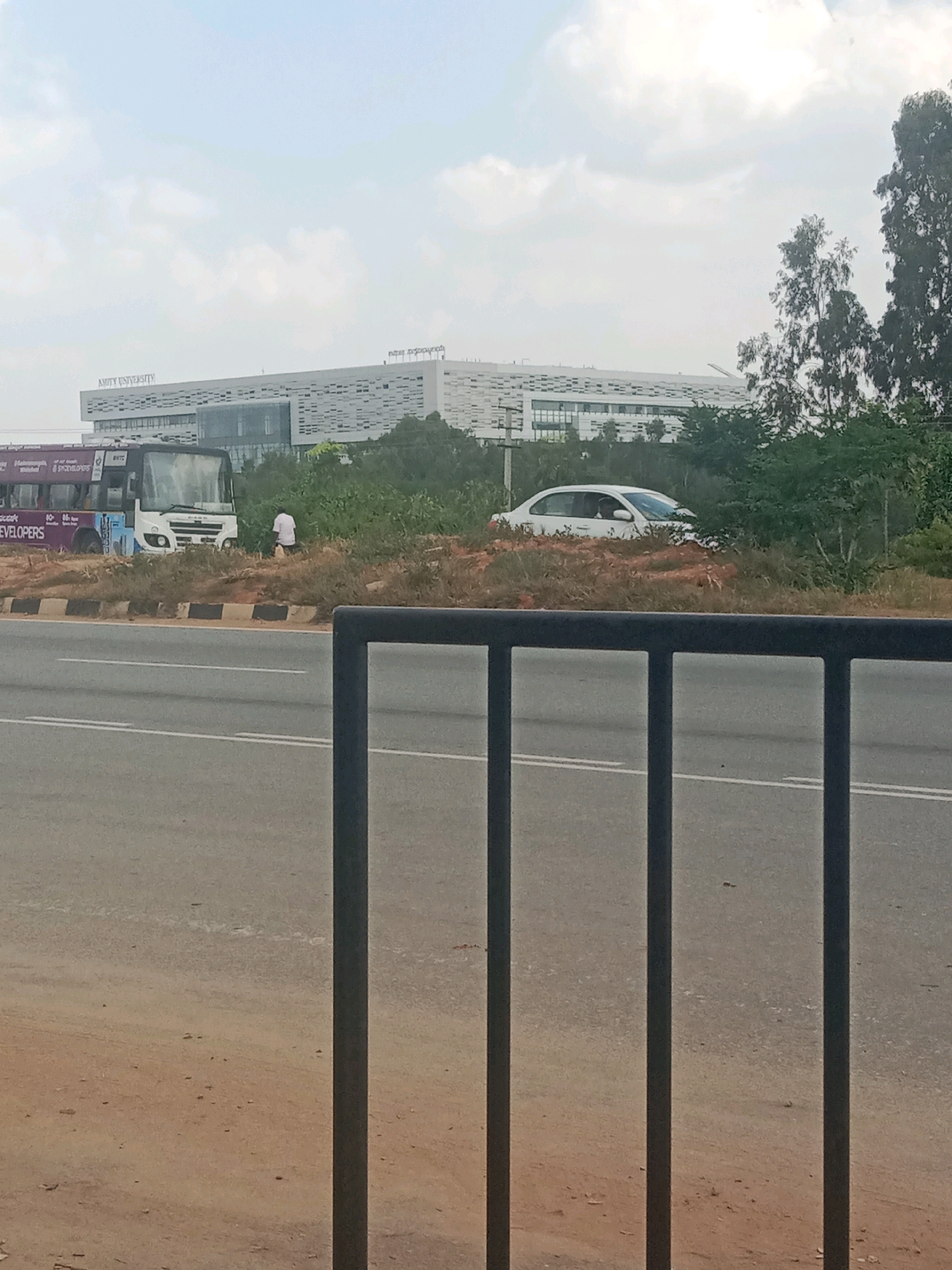 A wide-angle, eye-level, outdoor, daylight shot shows a modern, white building with an "AMITY UNIVERSITY" sign on its facade in the background. The building has a distinctive patterned facade and is surrounded by trees and vegetation. In the foreground, a highway runs horizontally across the frame, with a black metal fence in the immediate foreground. A white car is parked on the shoulder of the highway, facing away from the camera, partially obscured by foliage. To the left of the car, a white bus with purple and white graphics is stopped, and a person in a white shirt is standing near the bus and vegetation. The sky is overcast with light clouds. The location is Birasandra, India.