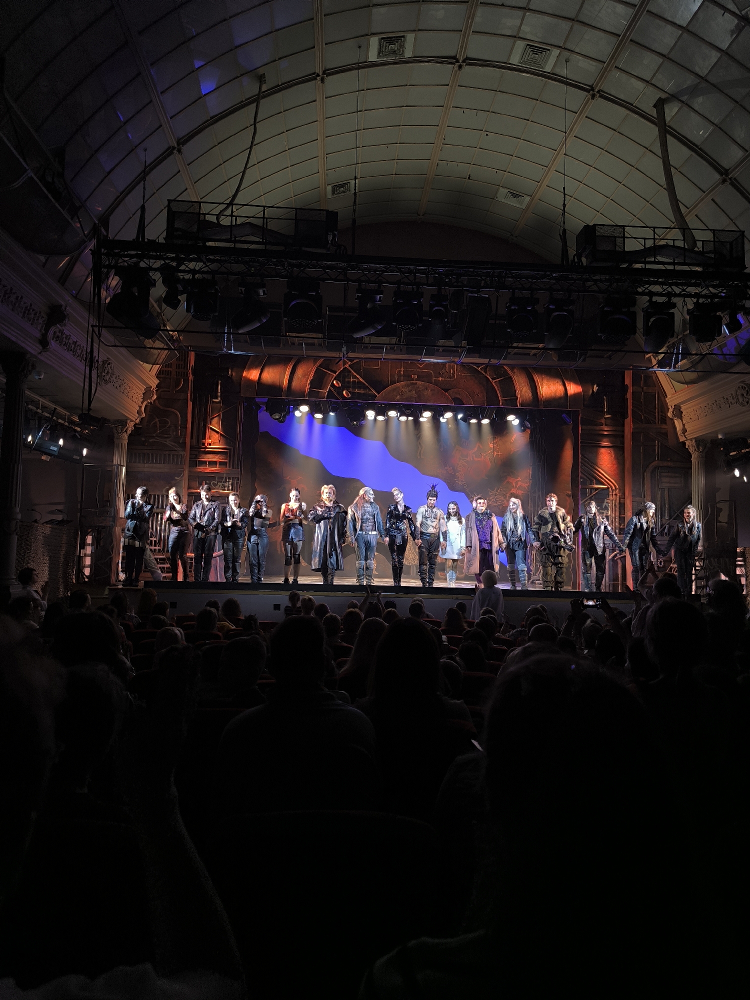 The image depicts a stage performance in a theater, with a group of actors on stage taking a bow. The stage is set with a rustic, industrial backdrop, featuring large, weathered structures and exposed metal elements. Several spotlights illuminate the actors and the stage. The actors are dressed in elaborate, possibly theatrical costumes, suggesting a dramatic or fantasy production. Some actors are wearing dark, leather-like attire, while others have more ornate and possibly historical-looking outfits. The audience is visible in the foreground, mostly in silhouette due to the lighting, with some audience members holding up phones, likely to record or photograph the performance. The theater itself appears to be an older building, with a high, arched ceiling and ornate architectural details on the sides, suggestive of a historical venue. The overall atmosphere is one of culmination and appreciation, with the actors acknowledging the audience. The image is taken from a perspective within the audience, looking towards the stage.