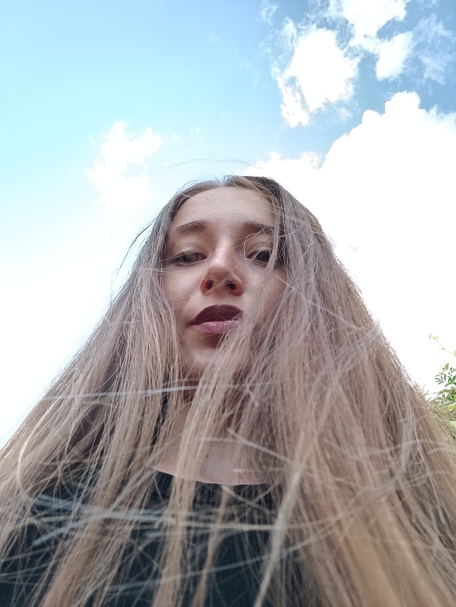 A low-angle, wide shot captures a young woman's face from a perspective looking up at her from below. Her long, blonde hair is a prominent feature, falling around her face and obscuring parts of her head and shoulders. She is wearing dark clothing, partially visible beneath her hair. The background is dominated by a bright blue sky with scattered white clouds, suggesting an outdoor setting. No specific location in Vypasne, Ukraine is depicted.