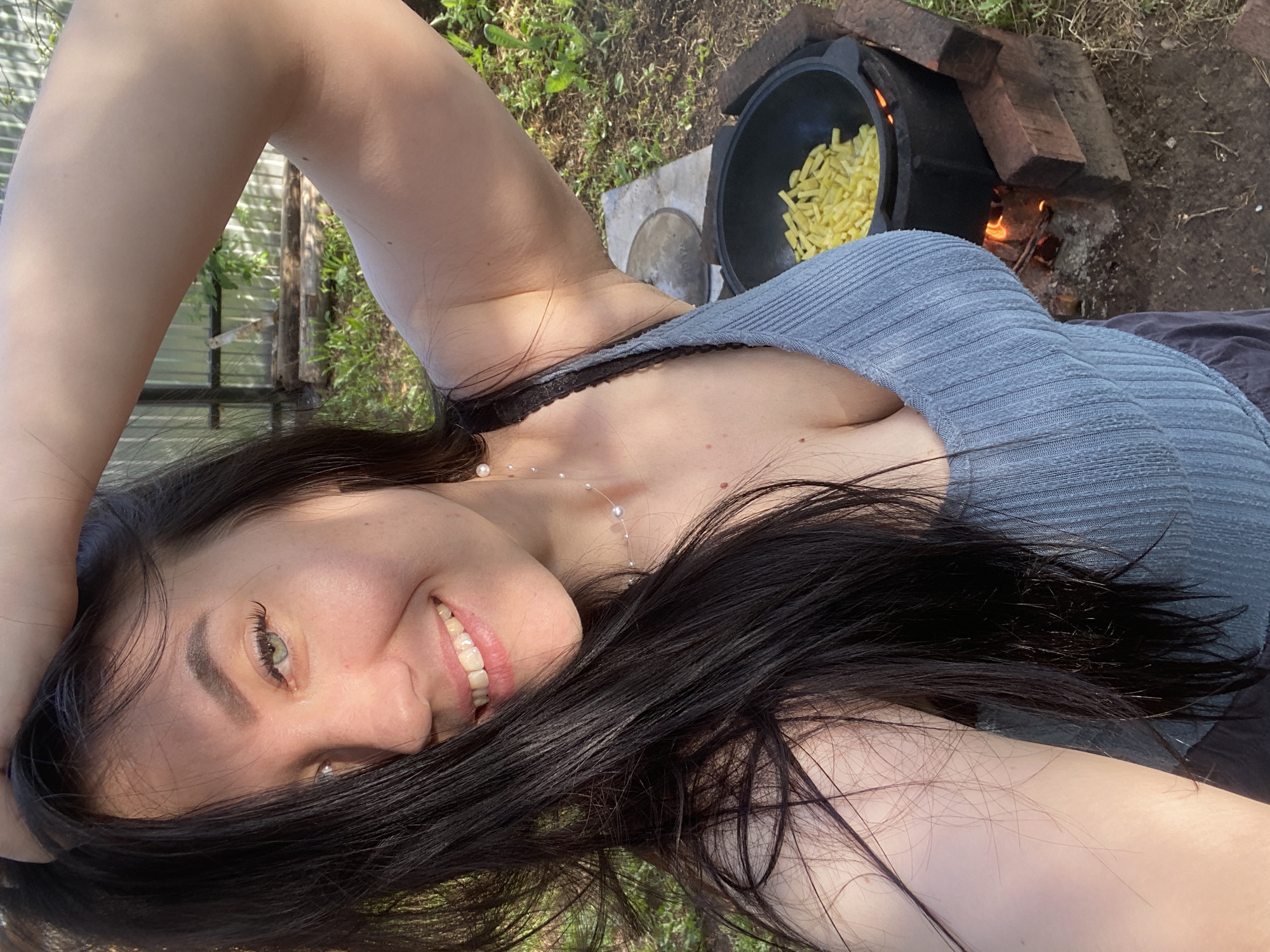 A woman is lying on the ground outdoors, smiling and looking at the camera. She has dark hair, fair skin, and is wearing a gray top and a pearl necklace. Beside her is a black cauldron filled with yellow food, possibly French fries, sitting over a wood fire contained by bricks. The ground appears to be covered in grass and dirt. In the background, there is a blurred view of some kind of structure, possibly a shed or greenhouse, suggesting a yard or garden setting. This photograph was taken in Russia, city of Chaikovsky.