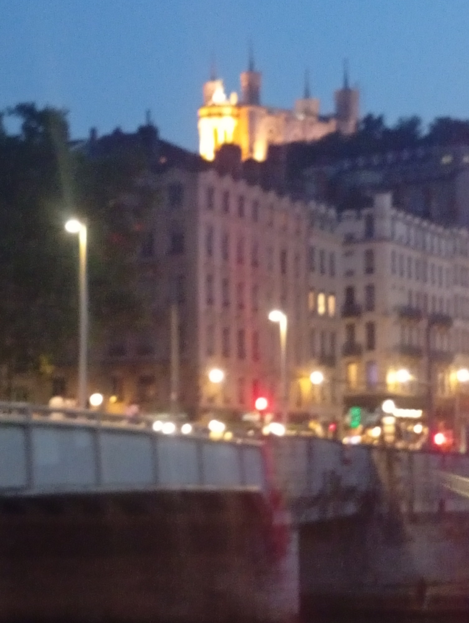 The image depicts a nighttime scene in Lyon, France, likely from a bridge overlooking the Saône or Rhône River. In the background, the Basilica of Notre-Dame de Fourvière is illuminated, its prominent towers and ornate architecture silhouetted against the deep blue twilight sky. The foreground features a section of a bridge, possibly Pont Kitchener-République or Pont de l'Université, with streetlights and building lights creating a warm glow. Buildings line the riverbanks, their windows displaying a variety of illuminated squares. The overall impression is a softly focused, atmospheric view of Lyon at dusk.