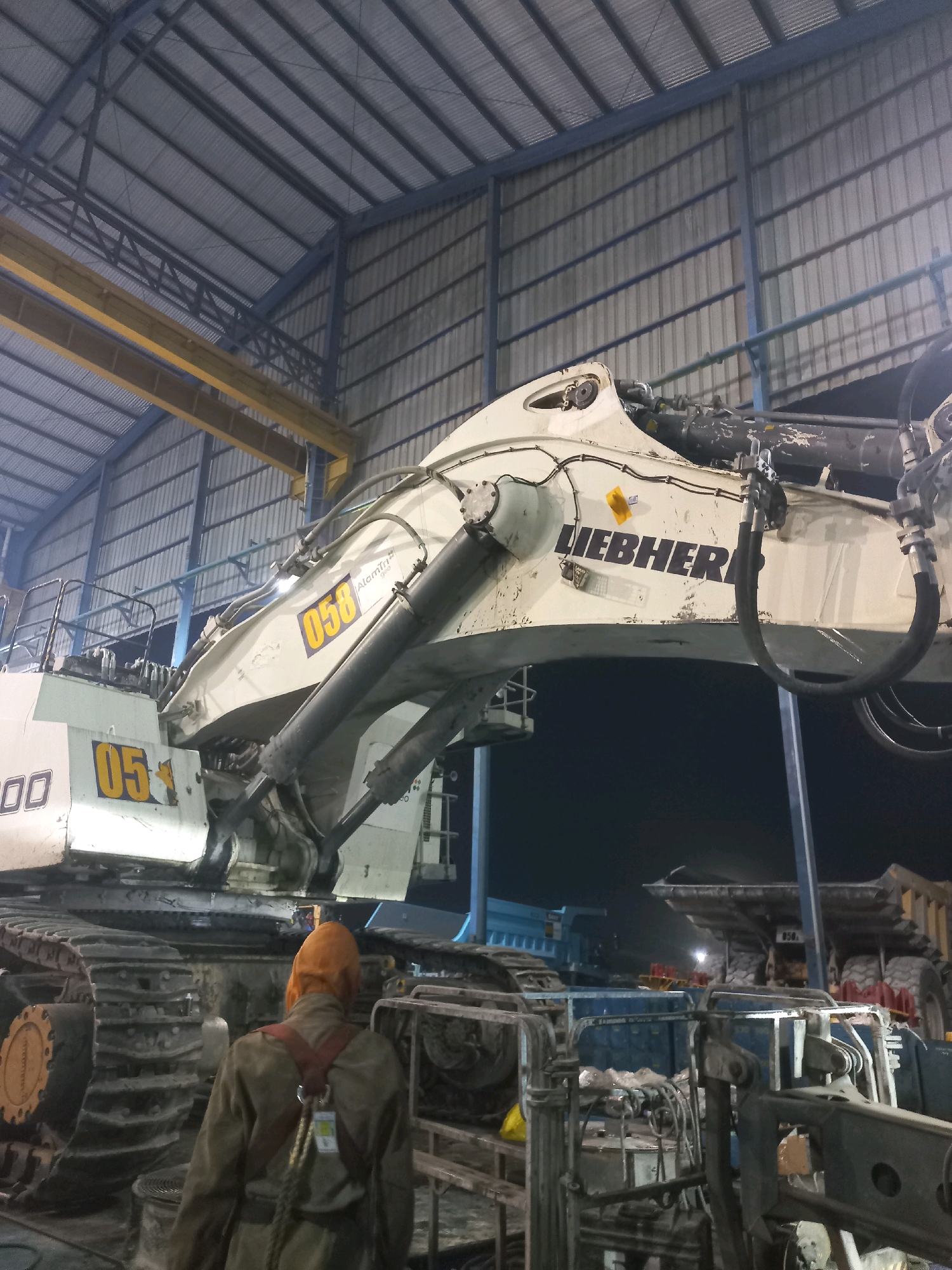 The image depicts a large, white Liebherr excavator in an indoor industrial setting, likely a workshop or storage facility, based on the corrugated metal walls and roof. The excavator is massive, with visible tracks and hydraulic components. The brand name "LIEBHERR" is clearly visible on the side of the excavator's boom. Two yellow numerical decals, "058" and "0500", are present on the excavator's body. In the foreground, a person wearing an orange head covering and a brown jumpsuit with a safety harness is visible from behind, facing away from the camera and towards the excavator. The environment appears to be dimly lit, suggesting it might be nighttime or that the facility's lighting is not very bright. In the background, other large mining or construction vehicles, including dump trucks, are partially visible. The presence of these heavy-duty machines, along with the industrial setting and a worker in safety gear, suggests this scene is related to heavy machinery maintenance or operation, potentially in a mining context. The specific location in Paringin, Indonesia, implies this is likely a facility associated with mining operations in that region.