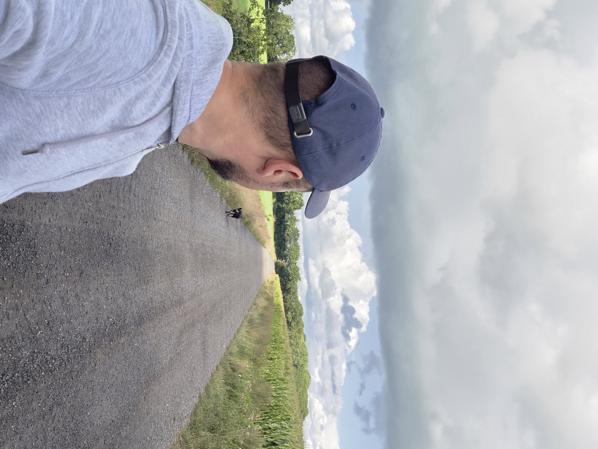 The image shows a man wearing a blue baseball cap and a grey hooded jacket, seen from behind, looking down a gravel road. A small black and tan dog is walking on the road ahead of him. The road is bordered by fields of green corn on the right and grass and trees on the left. The sky is cloudy with some patches of blue. The location is identified as Schiffdorf, Germany.