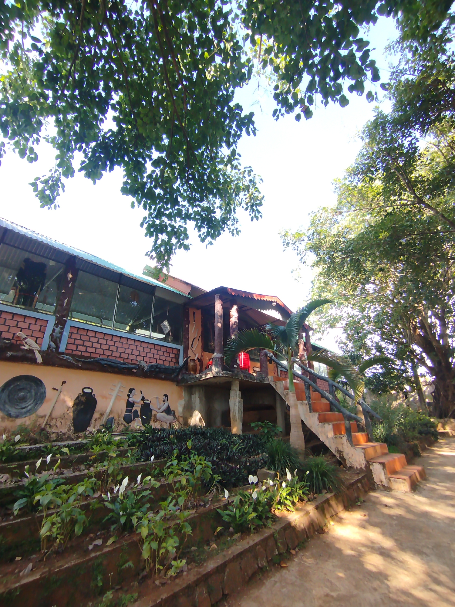 The image shows a building with a brick and stucco exterior, located in An Phu Ward, Vietnam. A covered patio area is on the left, enclosed with large windows. The stucco section features a mural depicting people and objects, including pots and musical instruments. A set of brick stairs with a log railing leads up to the patio. In front of the building are terraced gardens with flowering plants. Trees overhang the building, casting shadows. The ground is dirt and sunlight is visible.