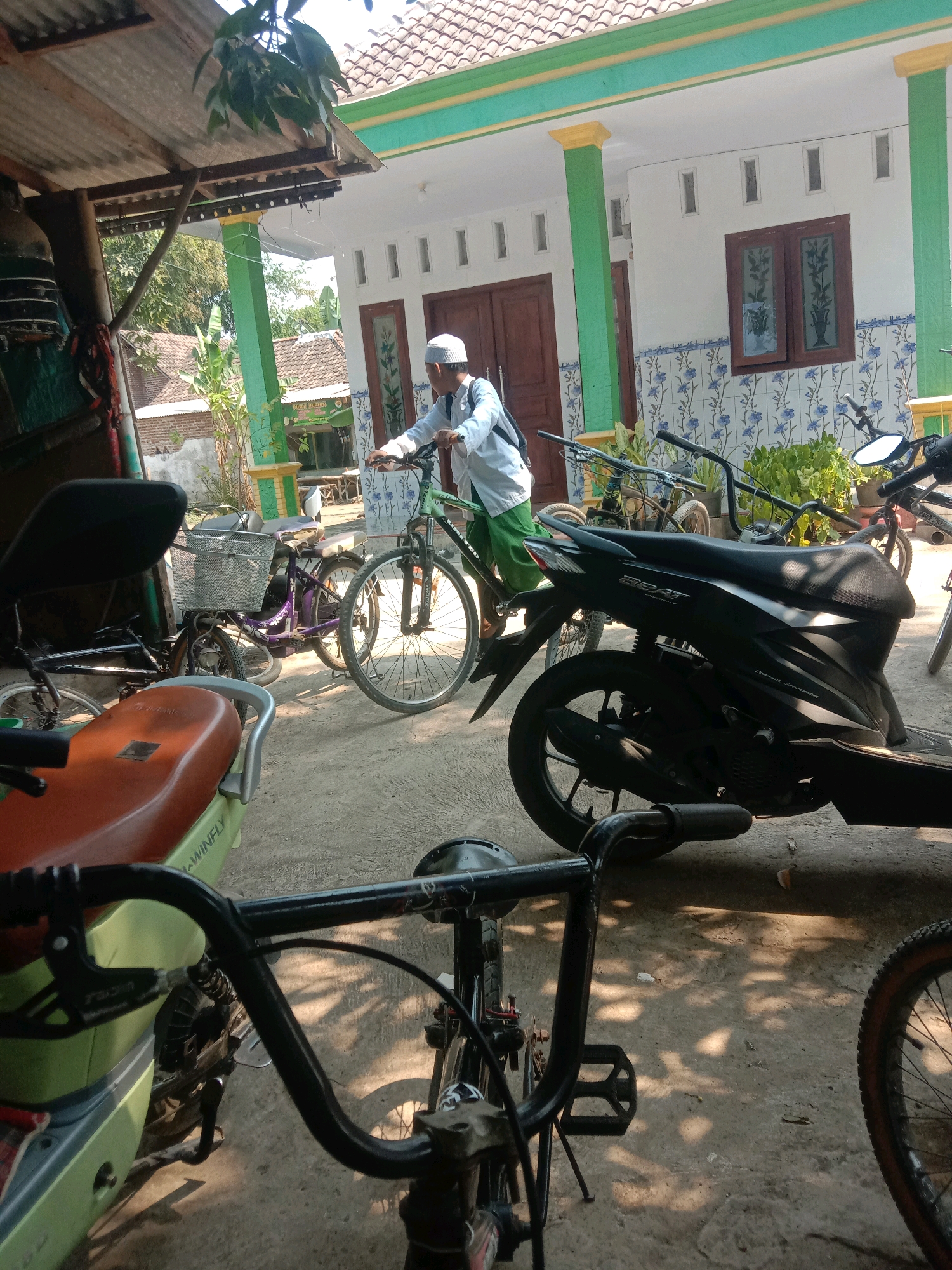 In Ngempit, Indonesia, a man wearing a white skullcap and a light-colored long-sleeved shirt with green pants stands next to a bicycle, holding its handlebars. The bicycle is dark green. Behind the man is a white building with green and yellow trim on its pillars and roofline. The building has two brown wooden doors and a wooden-framed window with intricate glass panes. A tiled wall with a blue floral pattern decorates the lower section of the building. Several other bicycles and a black scooter are parked in front of the building. The ground is concrete, and sunlight casts shadows from overhead foliage and structures. The scene appears to be an outdoor area, possibly a yard or pathway in front of a residence or community building.