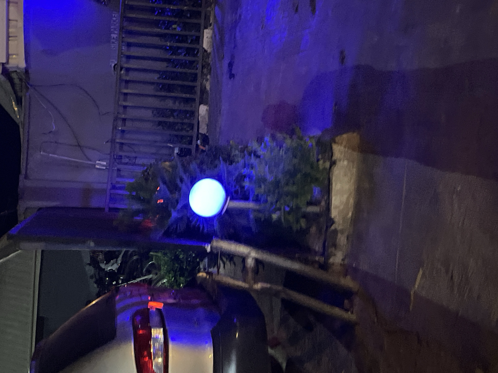 A nighttime shot focuses on the back of a white vehicle parked on a street in Akwanga, Nigeria. The vehicle, positioned at an angle, displays its taillights illuminated. Above the vehicle, a round, blue glowing orb light stands out against the darkness, positioned amidst foliage. In the background, a metal structure resembling a staircase can be seen, along with a wall. The overall scene is dimly lit, with a blue tint permeating the image.