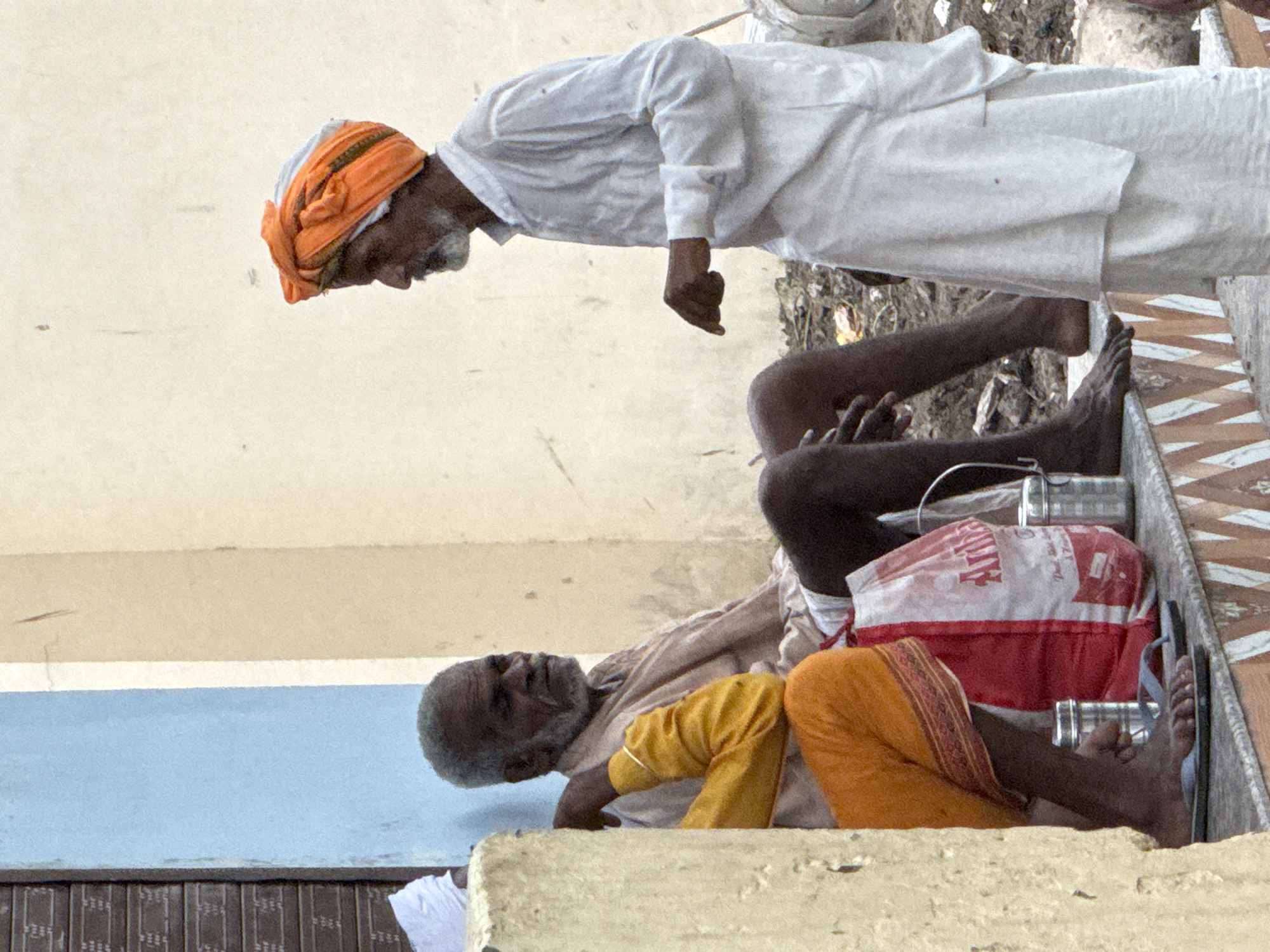 Two men are sitting on steps outdoors. One man, on the left, is older with graying hair and beard. He is wearing a light-colored shirt and white bottoms. He is looking to his right and his left arm is resting on his knee. In front of him are two metal containers and a red and white patterned bag. His legs are bent at the knees.
The second man, on the right, is wearing a white shirt and trousers, and an orange and white turban. He is facing the seated man and appears to be speaking to him, with his right hand extended slightly. The background consists of a light-colored wall and some debris on the ground. The setting appears to be on a street or public area in Rishikesh, India, given the attire and architectural style.