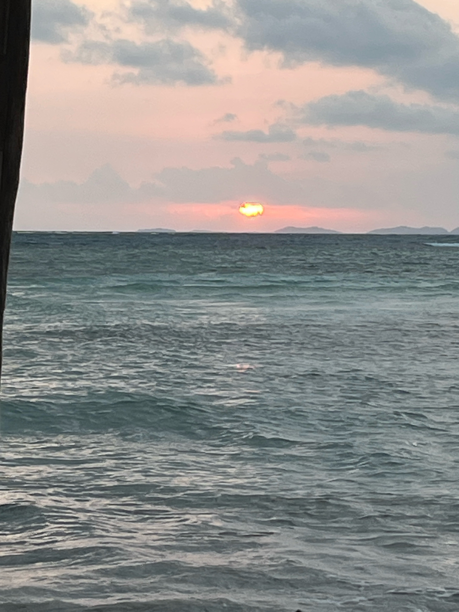 The image depicts a tranquil scene of the ocean at sunset, likely taken from a beach or pier in Karimunjawa, Indonesia. The sky is painted with soft hues of pink and orange, with scattered clouds adding texture. The sun, partially obscured by clouds, casts a warm glow on the horizon, its reflection shimmering on the gently rippling water. In the distance, a faint silhouette of islands can be seen. The foreground is dominated by the vast expanse of the ocean, with its surface creating a mesmerizing pattern of waves. A dark vertical element on the left edge of the frame suggests a wooden structure, perhaps part of a pier or a building. The overall atmosphere is serene and peaceful, capturing the natural beauty of a tropical sunset.