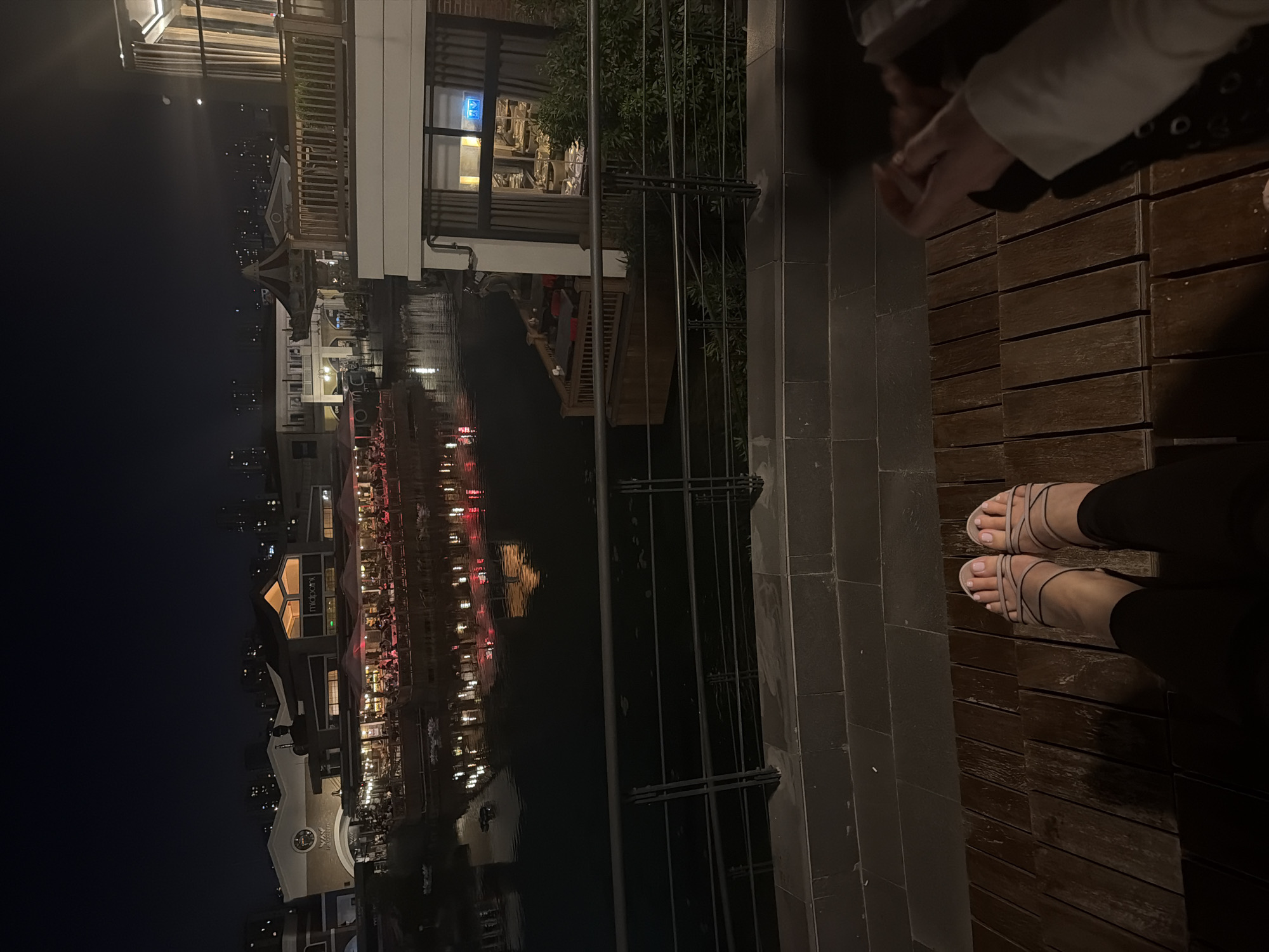 An image depicts a nighttime scene at an outdoor location. On the right side of the image, two individuals are visible, one with feet in sandals. A dark river reflects lights from buildings, restaurants, and possibly other illuminated structures on the left side. Based on the image's context, the location may be in Küçükçekmece, Turkey.