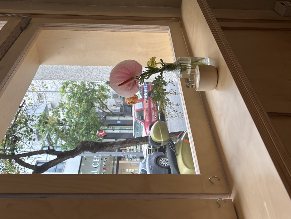 The image depicts an urban street scene in Tel-Aviv, Israel, viewed from inside through a wooden-framed window.
In the foreground, a light-colored plywood window frame surrounds the view. A matching wooden windowsill extends across the lower portion of the frame. On this windowsill, a light beige ceramic mug containing a beverage with latte art is positioned to the left. To its right, a clear, ridged glass vase holds a small floral arrangement consisting of a prominent pink anthurium, a smaller orange flower, a yellow flower, and green foliage with small berries. Two silver-colored metal hooks are attached to the lower left vertical window frame. The interior wall to the right of the window appears light-colored and textured.
Through the window, an urban street is visible under natural light. Parked vehicles include a partially obscured dark grey car on the left, an olive-green scooter (Vespa-style) in the center with its mirrors reflecting, and a red pickup truck on the right. The pickup truck features white graphics and Hebrew text on its side, including "קארגו" (Cargo) and a partial phone number. Behind the vehicles, commercial buildings line the street. A prominent storefront on the left displays "AL CEN" and Hebrew text "ראייה • שו" (Ra'aya • Sho), suggesting an optical center. Several trees with varying foliage are distributed along the street, one with a prominent trunk in the center-left. A red circular street sign with white elements is visible above the red pickup truck. No individuals are visible.