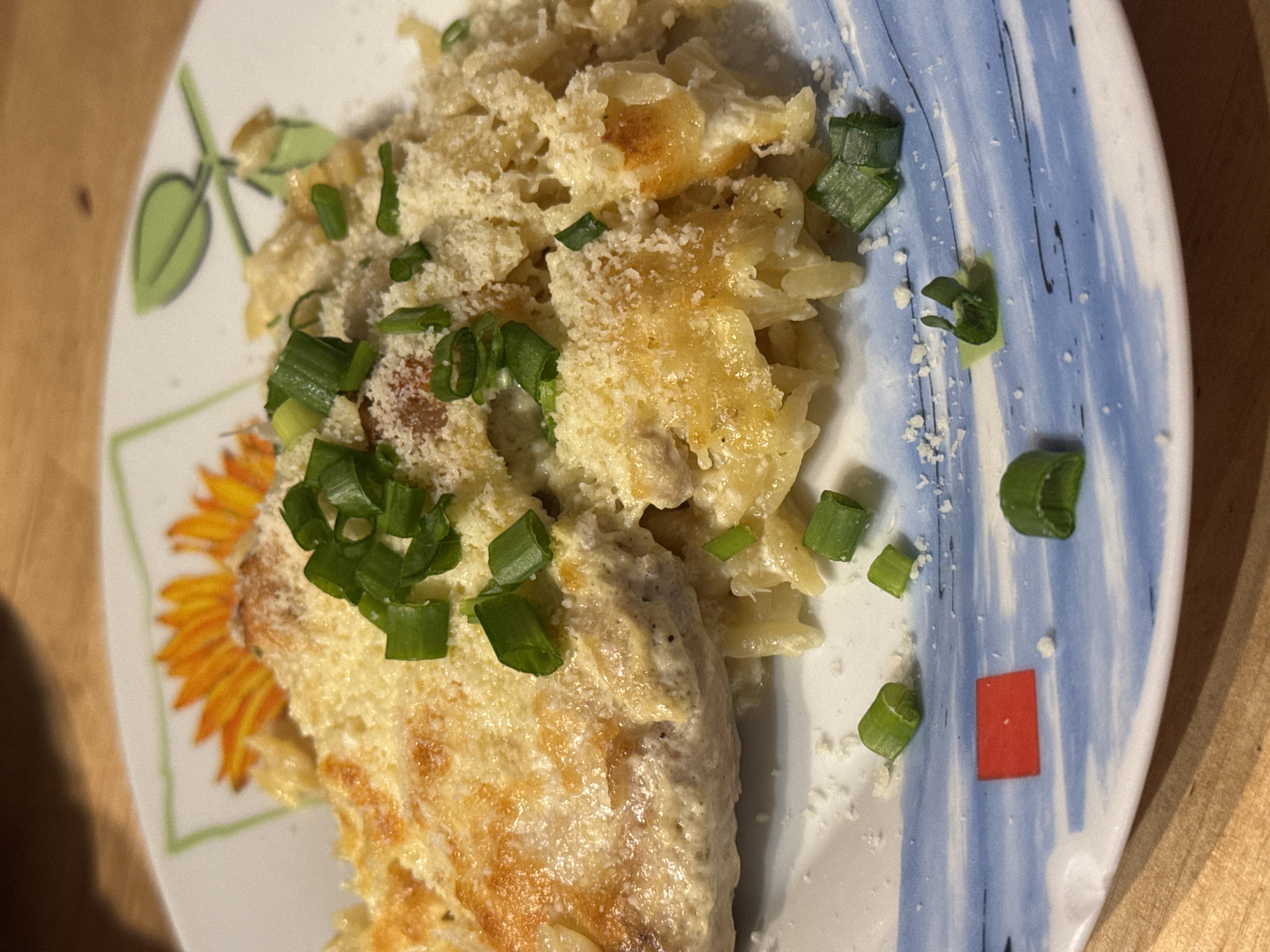 A close-up shot depicts a plate of food. The dish includes a portion of pasta, possibly orzo, covered in a creamy sauce and topped with chopped green onions and grated cheese. Additionally, there is a piece of chicken, also covered in the same creamy sauce and garnished with green onions. The plate has a design incorporating a sunflower and blue stripes. A small red square is also on the plate. It is not possible to determine the location of the city of Sofia, Bulgaria from the image.