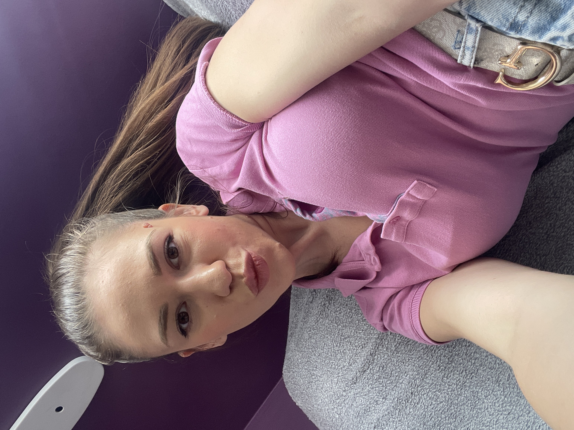 A woman is shown in a selfie-style shot from a slightly high angle. The woman has brown hair, fair skin, and is wearing a pink polo shirt and a designer belt with a gold buckle. She appears to be sitting or lying on a gray, textured couch or surface. The background includes a purple wall. There is no discernible information to pinpoint the specific location within Sofia, Bulgaria.