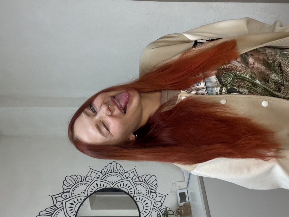 A young woman with long, straight auburn hair winks and sticks out her tongue at the camera. She has a nose ring and earrings. She is wearing a tan jacket with white sleeves and a graphic t-shirt. Behind her is a white wall with a black and white mandala design above a mirror. A small teddy bear, a thermostat, and some notecards sit to the right.