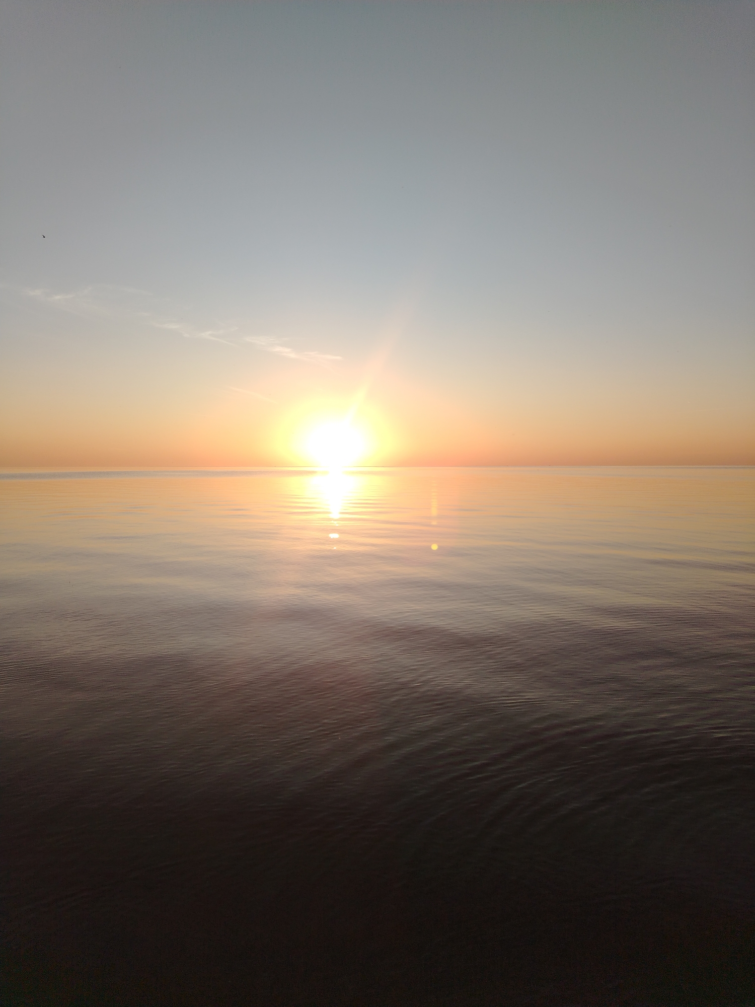 The image depicts a serene seascape at sunset or sunrise. The sun, a bright, luminous orb, is positioned near the horizon, casting a warm, golden glow across the sky and water. The sky transitions from a pale blue at the top to a soft orange and yellow near the horizon. A faint, wispy cloud is visible in the upper left quadrant. The surface of the water is calm, with subtle ripples reflecting the colors of the sky and the sun's radiant light. The lower portion of the image is darker, suggesting the water deepens or the light diminishes further from the sun. The location is identified as the city of Ivanovskoe, Russia. There are no people, man-made objects, or specific landmarks visible in this natural landscape scene.