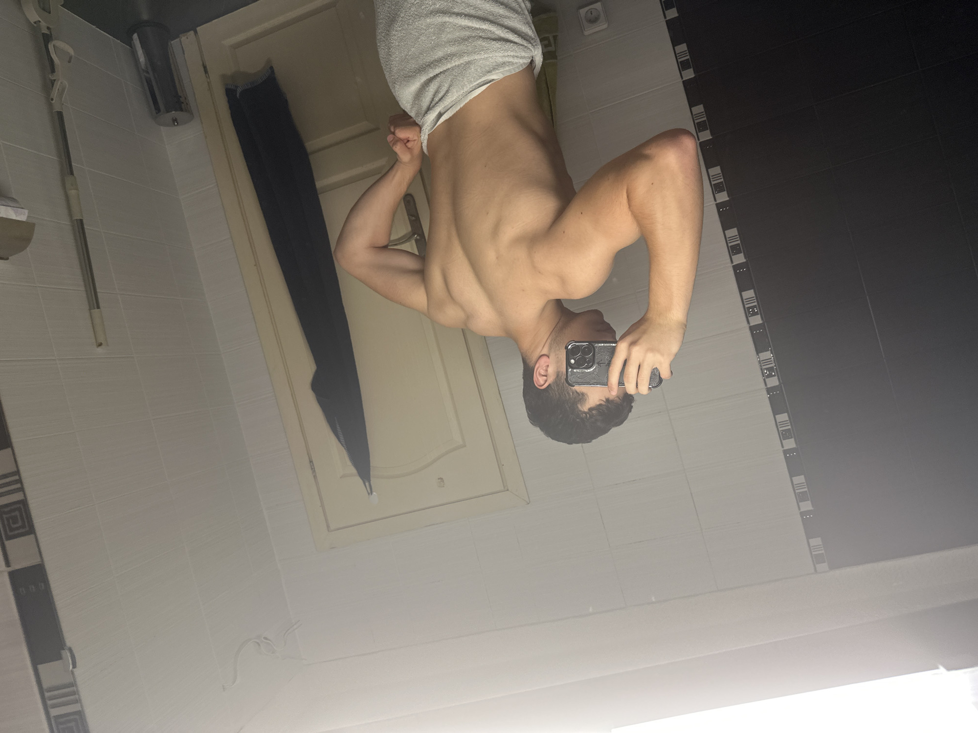 In a bathroom in the city of Unknown, Unknown, a young adult male is captured in a mirror selfie, facing away from the reflective surface and presenting his bare, muscular back. He has short, dark, curly hair and is wearing a white towel wrapped around his waist. His left arm is flexed, showcasing developed biceps and deltoids, while his right hand holds a dark-colored smartphone (with a patterned case) to take the photograph, obscuring his face. The reflection reveals a bathroom with predominantly white square tiles on the walls, segmented by a vertical strip of black and white geometric pattern tiles on the left and a lighter decorative strip with a swirling white motif on the upper right. The floor is dark-tiled. A bright overhead light source creates a glare at the top of the mirror reflection. Visible objects in the background include a light-colored wooden door with a metallic handle, from which a dark blue towel or garment hangs. A cylindrical metallic trash bin, a long-handled cleaning tool (possibly a mop), a white power outlet, and a toilet paper holder are also discernible in the reflection. A light green towel hangs on a hook on the wall behind the subject's reflected lower body.