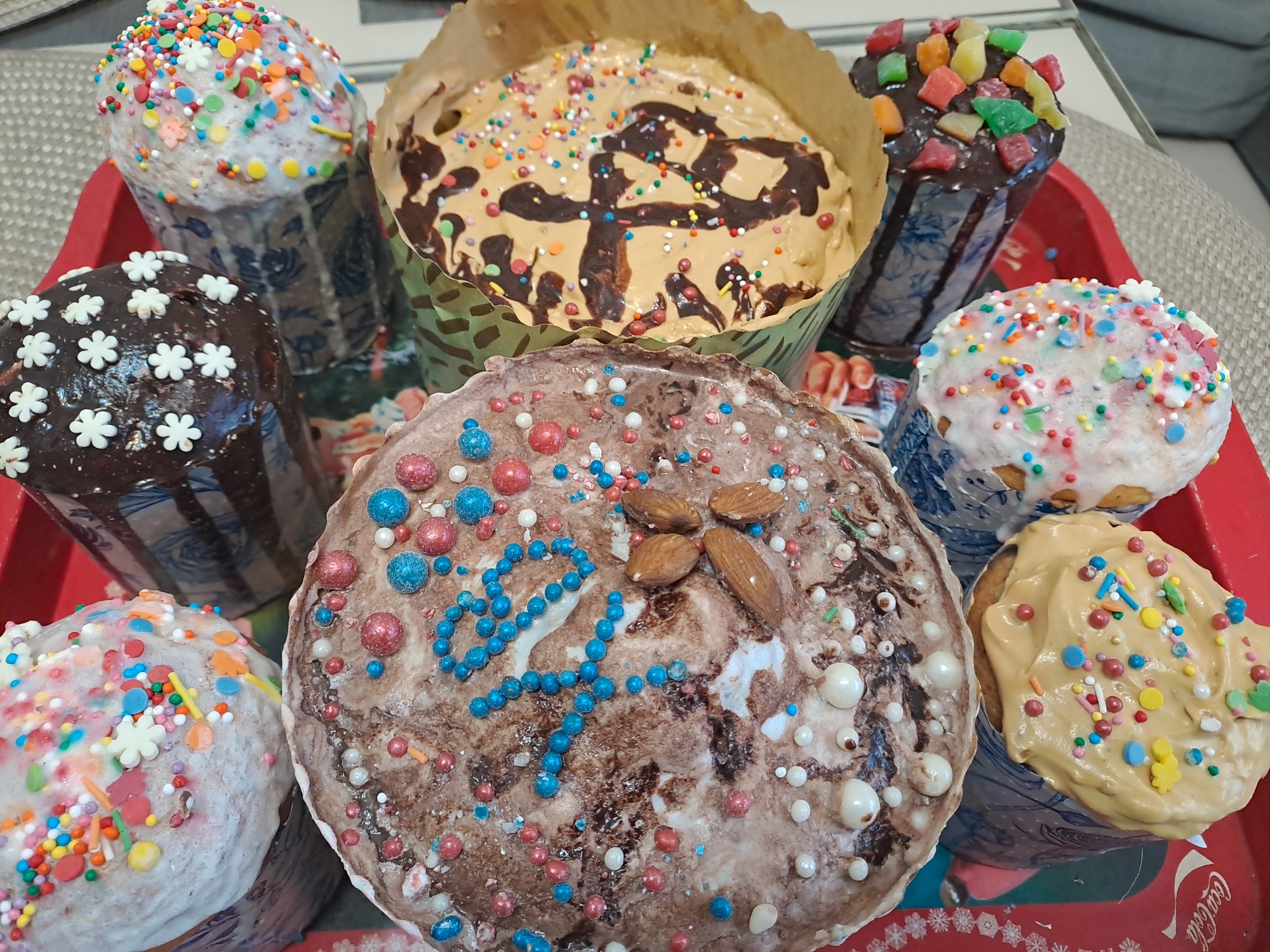 The image shows a collection of elaborately decorated Easter breads, a traditional food in Russia. They are arranged on a red tray with a Coca-Cola logo. The breads vary in size and decoration, featuring different types of frosting, sprinkles, candied fruits, and nuts. The location is likely a home in Elektrostal, Russia, based on the Russian Easter bread tradition. There are no people visible in the image.