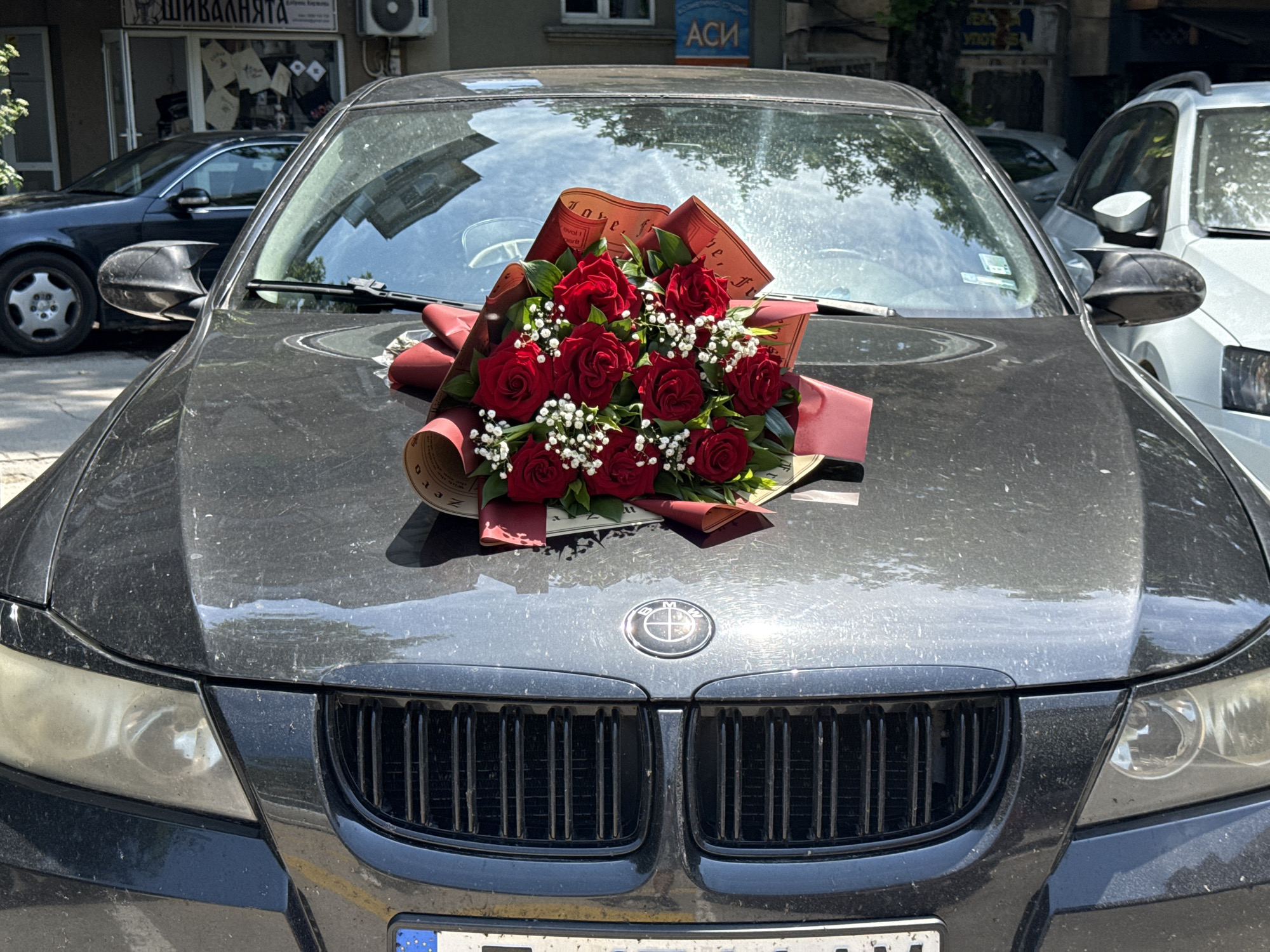 The image features a dark-colored BMW car, possibly black or dark gray, parked outdoors during what appears to be a sunny day. A prominent bouquet of vibrant red roses, interspersed with white baby's breath (Gypsophila) and green leaves, is resting on the car's dusty hood. The flowers are wrapped in decorative brown and red paper, which has some faint, handwritten-style text that includes words like "love" and "sweet."
The car's front grille is black, and the BMW emblem is clearly visible on the hood. The headlights appear somewhat oxidized or foggy. A partial view of a European-style license plate with a blue strip is seen at the bottom.
In the background, a building with a storefront is visible. The store features a sign with Cyrillic text that reads "ШИВАЛНЯТА," translating to "The Sewing Room" or "Tailor Shop." Another sign above it displays "АСИ" in blue letters. An air conditioning unit is attached to the building's exterior. Reflections in the car's windshield show parts of the surrounding environment, including other vehicles and greenery. The scene suggests a street setting, consistent with Varna, Bulgaria, given the Cyrillic text and license plate style. The presence of the rose bouquet on the car hints at a celebration or a special occasion.