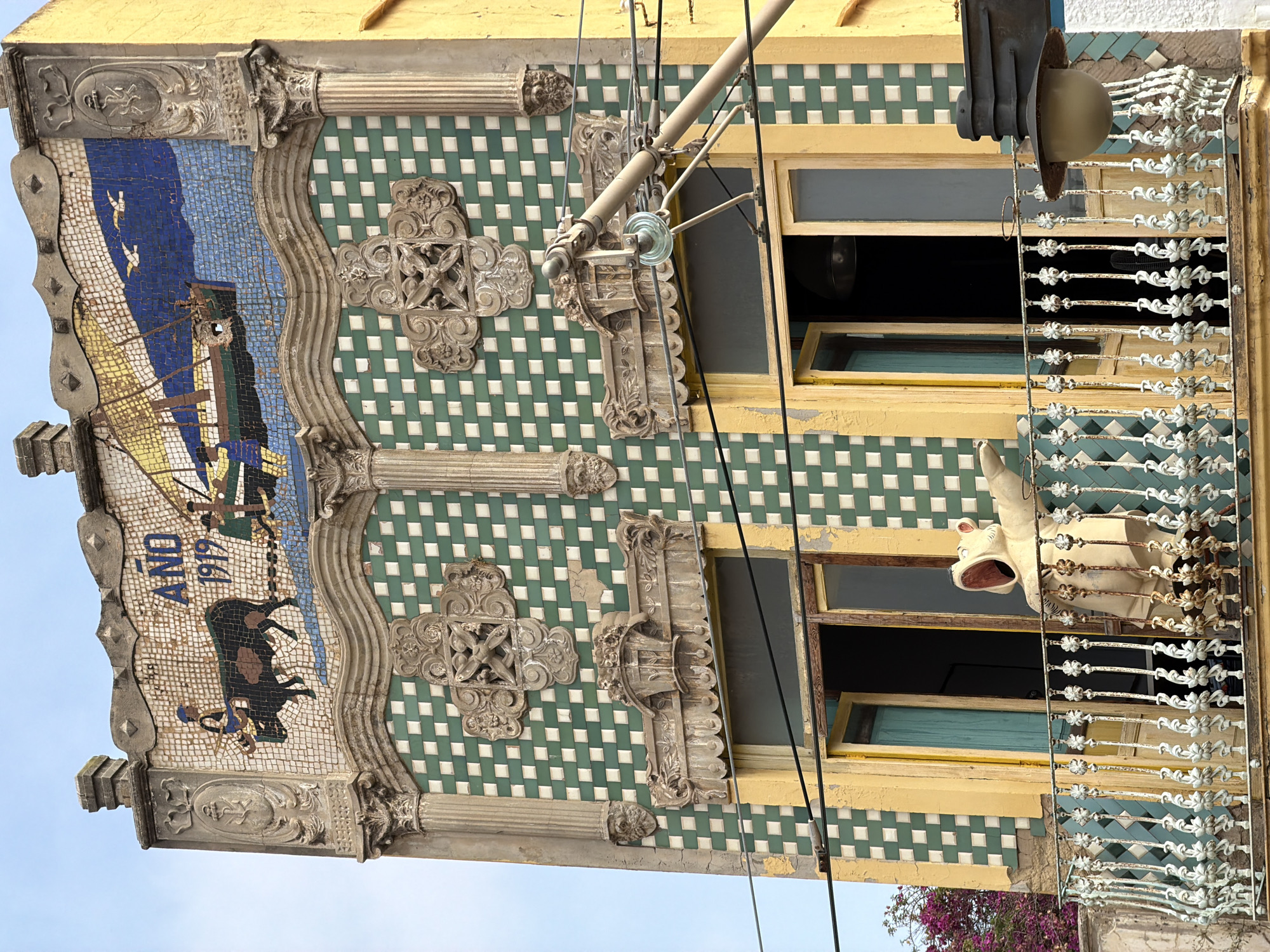A facade of an old building features a mosaic artwork at the top depicting a bull being led by a person towards a ship at sea, with two birds flying overhead. The year "1919" is also visible in the mosaic. The building's facade is decorated with green and white square tiles. Ornate architectural elements, including columns and decorative carvings, are present. Two open windows are visible on the second floor, with a balcony railing below. A light-colored, cartoonish figure with its mouth open stands on the balcony, facing to the right. Overhead wires and a street lamp are also visible. The sky is a pale blue.