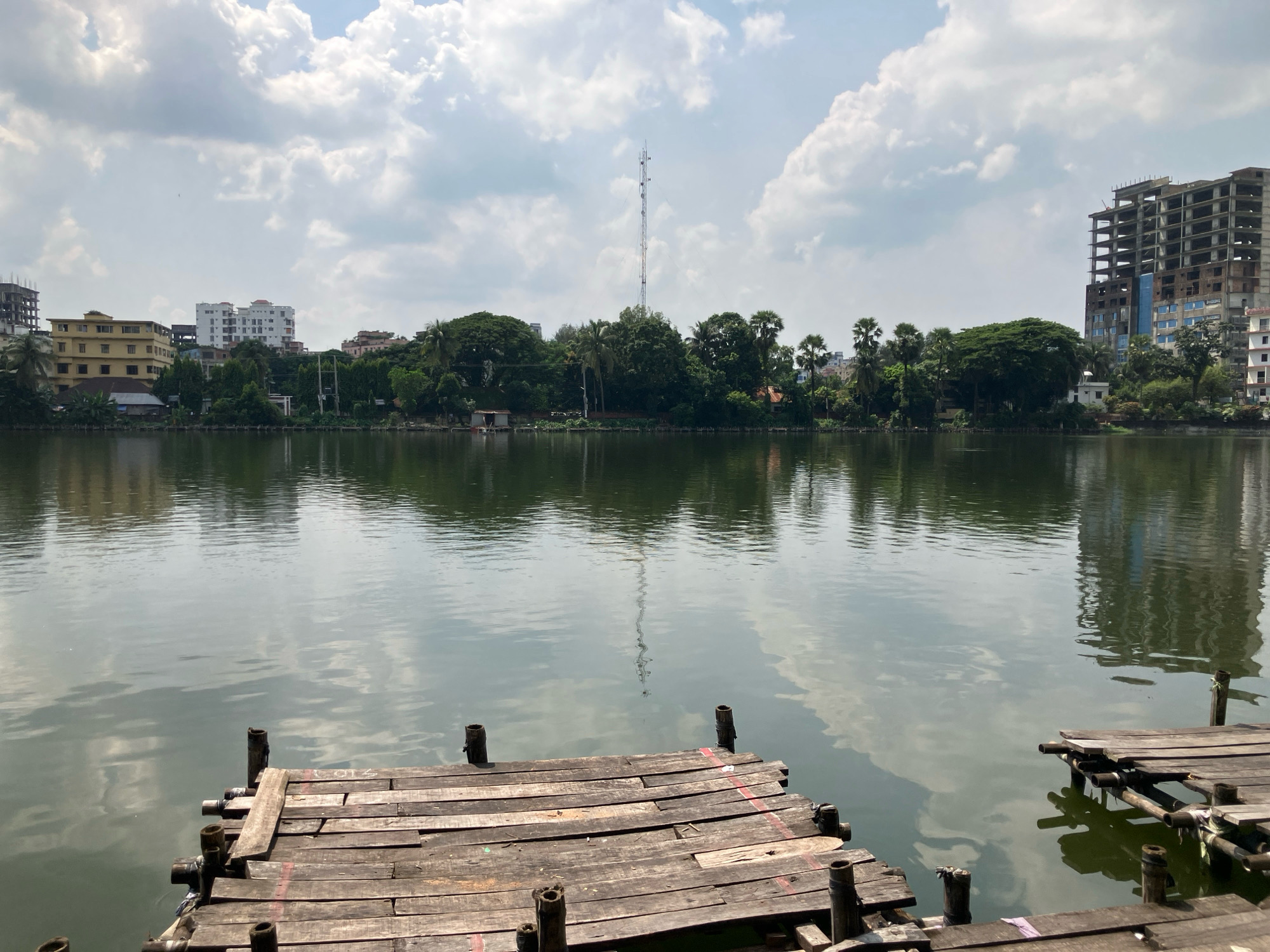 The image depicts a tranquil water body in Feni, Bangladesh, on a cloudy day. In the foreground, a weathered wooden jetty extends into the water, constructed from planks of varying sizes and shades of brown, supported by rough wooden posts. The water is calm, reflecting the sky and the distant cityscape. Across the water, a line of lush green trees forms a backdrop to several buildings.
On the left side of the scene, a yellow, multi-story building stands out, with other modern buildings visible behind it. Towards the center, a tall communication tower pierces the sky, surrounded by dense foliage. On the right, a partially constructed building, a skeletal structure of concrete and steel, dominates the skyline, with a completed modern building adjacent to it. The sky is filled with fluffy white clouds, casting soft shadows on the water. The overall impression is one of urban development juxtaposed with natural scenery.