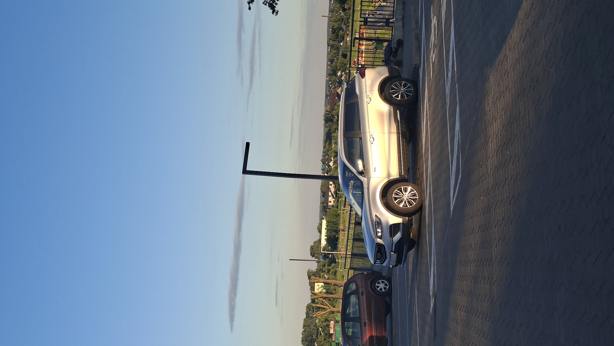 The image captures a parking lot scene in Smaliavichy, Belarus, under a clear blue sky with scattered clouds. A silver Geely SUV is parked in the foreground, facing right, with its driver's side visible. Its license plate, though partially obscured, shows characters resembling "BE625 OT". Adjacent to the SUV, a dark red minivan is parked on the left.
The parking lot surface is paved with dark grey interlocking bricks, marked with white parking lines. A black, L-shaped pole stands behind the silver SUV. In the background, a low black metal fence separates the parking lot from a grassy area with trees and buildings, indicative of a residential area. A few small, dark objects, possibly electric scooters, are visible near the fence behind the SUV. The overall lighting suggests it is late afternoon or early evening, with the sun casting long shadows on the parking lot. The horizon shows a scattering of houses and trees, typical of a town or suburban setting.