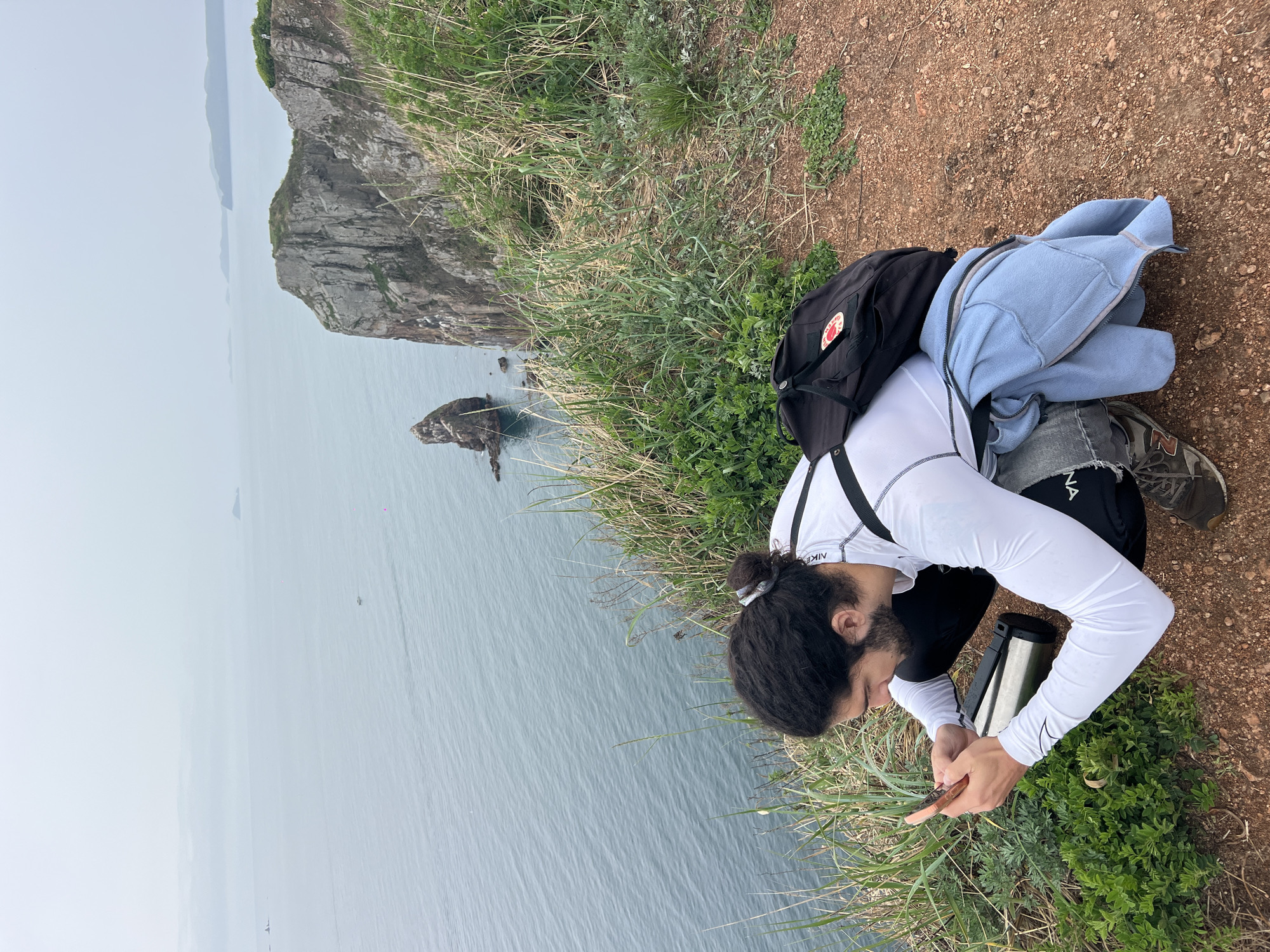 A man is crouched on a grassy, earthen cliff edge in Russkiy Island, Russia, overlooking the sea. He is focused on his mobile phone, his dark hair pulled back in a bun. He wears a white long-sleeved shirt, dark pants, and a black backpack. A light blue jacket lies near him. In the background, a rocky cliff juts out into the water. The sea is grey and misty. A metal container sits beside him.