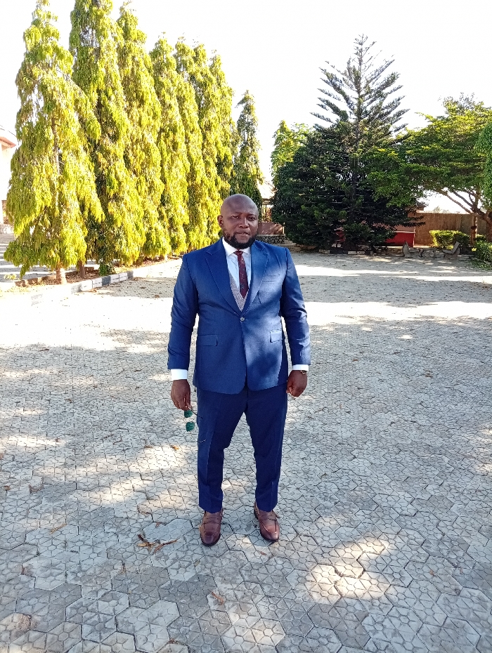 A man in a tailored navy suit, burgundy tie, and brown shoes stands on patterned paving stones. He is bald with a dark complexion and holds dark sunglasses in his left hand. Behind him are several tall, slender trees with dense foliage that glow yellowish in the sunlight. A larger, darker green tree is positioned to the right, with hints of buildings visible beyond it. The setting appears to be outdoors on a bright, sunny day.