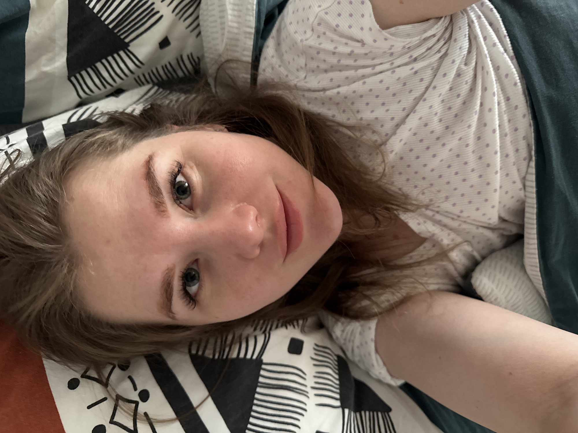A light-skinned female individual is captured in a close-up selfie, lying on a bed in Gornyi (Dvurechenskoe S/P), Russia. She has light brown hair, with visible facial features including green eyes and a slight smile. Her right arm is extended, likely holding the camera, partially visible on the lower right. She is wearing a white or light-colored, short-sleeved top adorned with small, light purple polka dots. Her head rests on a pillow with a black and white geometric pattern. A teal or dark green duvet or sheet covers her lower body and the bottom portion of the frame. The scene is illuminated by an unseen light source, casting subtle shadows.