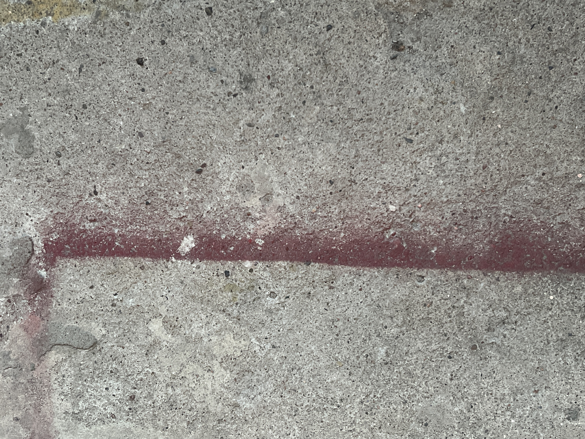 The image displays a close-up, top-down view of a rough, grey concrete surface with a vertical red spray-painted line running down the left side. The concrete has a speckled texture with visible aggregate. The red paint appears somewhat faded and uneven. The overall impression is of an outdoor urban surface, likely a sidewalk or wall, with graffiti. There are no people or other distinguishing objects present in the frame. The scene is static and devoid of any action.