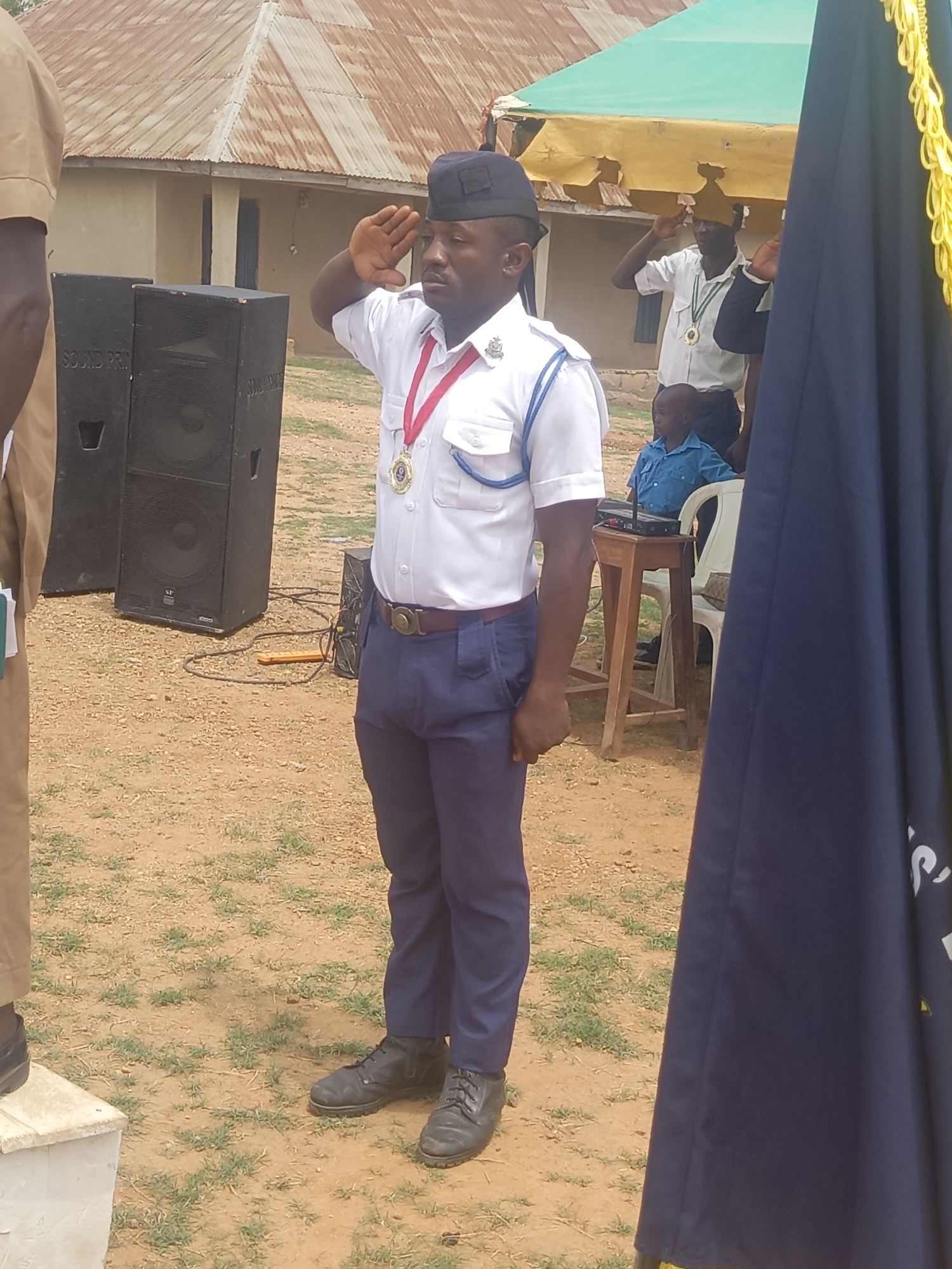 Akwanga, Nigeria: A man in a white shirt, blue pants, and black shoes salutes. He wears a red lanyard with a gold medal. He stands on a dirt surface with sparse grass. To his left are two black speakers. Behind him, partially obscured, is a man in a similar uniform. Behind that man, a child sits on a chair at a table. Overhead is a green and yellow tarp. To the right, partially obscuring the scene, is a navy blue flag with gold fringe. In the background is a building with a corrugated metal roof.