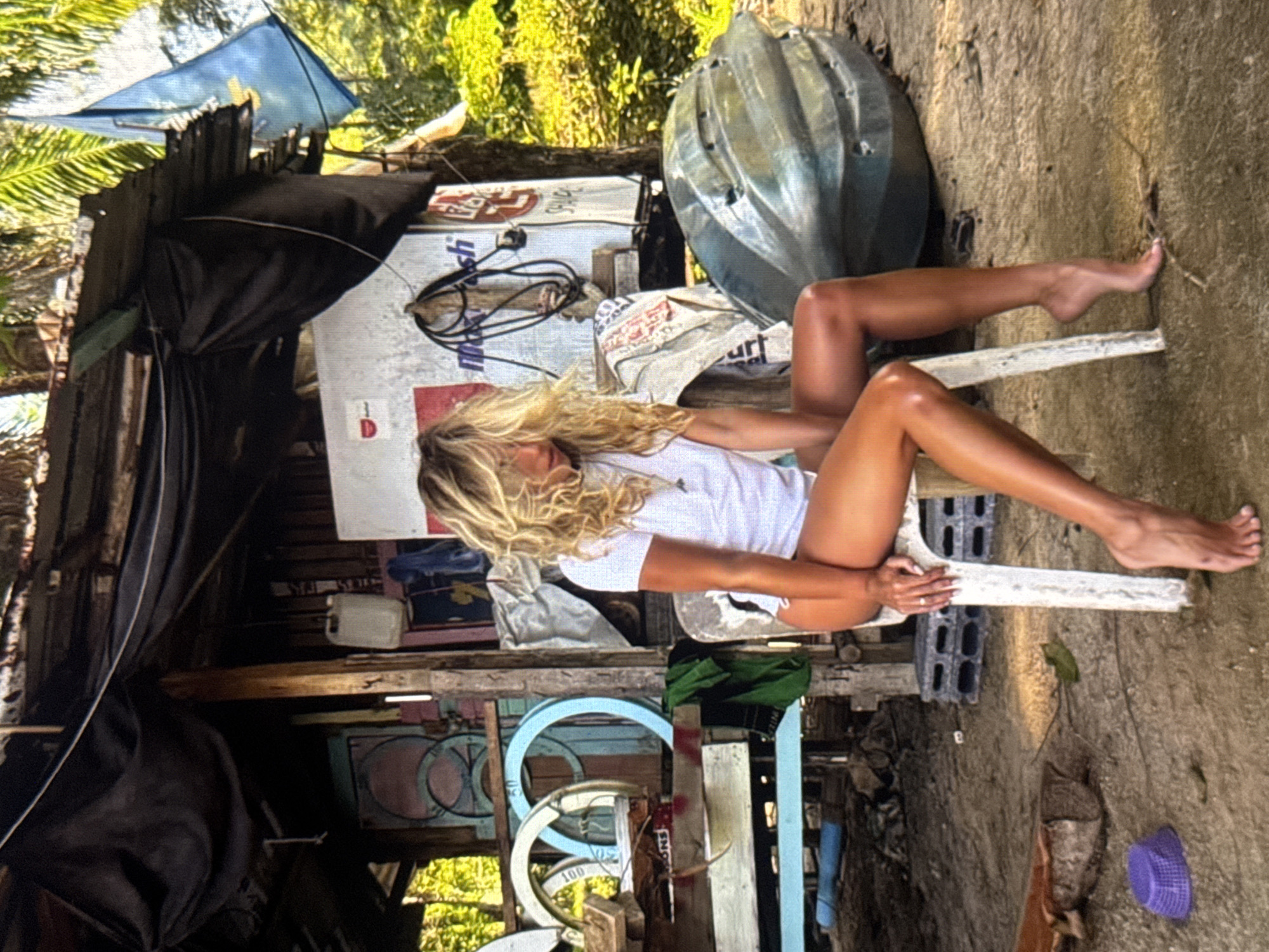 This media file depicts a woman with blonde hair sitting on a white plastic chair outdoors. She is wearing a white t-shirt and her legs are extended. The setting appears to be somewhat rustic, with weathered wooden structures and scattered objects. In the background, there are trees and foliage, suggesting a tropical environment. A large, rounded, green object, possibly a water tank or similar container, is visible to the right of the woman. Various items, including what looks like tarps, wires, and possibly building materials, are scattered around the area, contributing to a sense of disarray. A small purple object, resembling a basket or container, rests on the ground in the lower right corner.
The lighting suggests it is daytime, with sunlight filtering through the trees. The overall impression of the scene is casual and relaxed. There are no overt indications of a specific event or strong emotions.
Visible text includes what appears to be logos or branding on white material in the background, though it is difficult to read clearly. Some text, "MitchFsh" and "urt", is partially visible. There is also a red "D" shape on a white background.