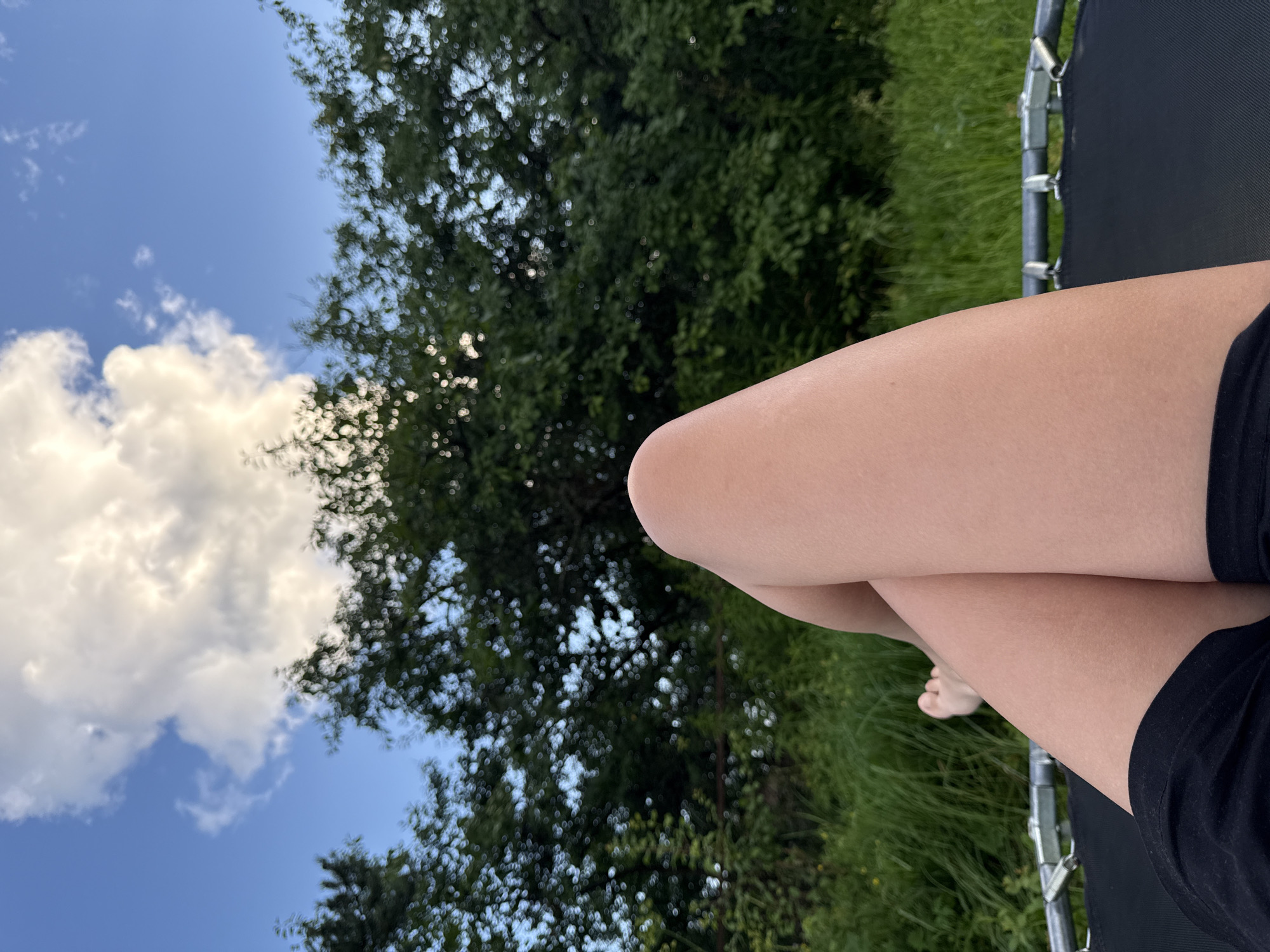 The image shows a close-up perspective from a person lying on a trampoline, with their legs crossed and visible in the foreground. The person is wearing black shorts. Behind the legs, a lush green grassy area and a dense line of trees are visible. The sky above is blue with a large, fluffy white cloud. The scene appears to be outdoors, likely in a backyard or park, during daylight hours. The overall impression is one of relaxation and enjoyment of a summer day. No specific landmarks or architectural features are visible to pinpoint the exact location within Vetluzhsky, Russia.