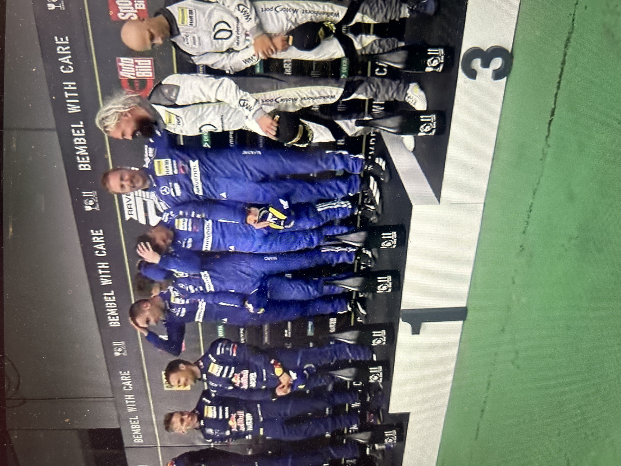 The image shows a group of people standing on a podium with numbers 1, 2, and 3, likely celebrating a victory. The individuals on the podium are wearing racing suits. There are team names or sponsor logos visible on their clothing. The backdrop includes text "BEMBEL WITH CARE." The image does not contain information that allows it to be placed in the city of Sofia, Bulgaria.