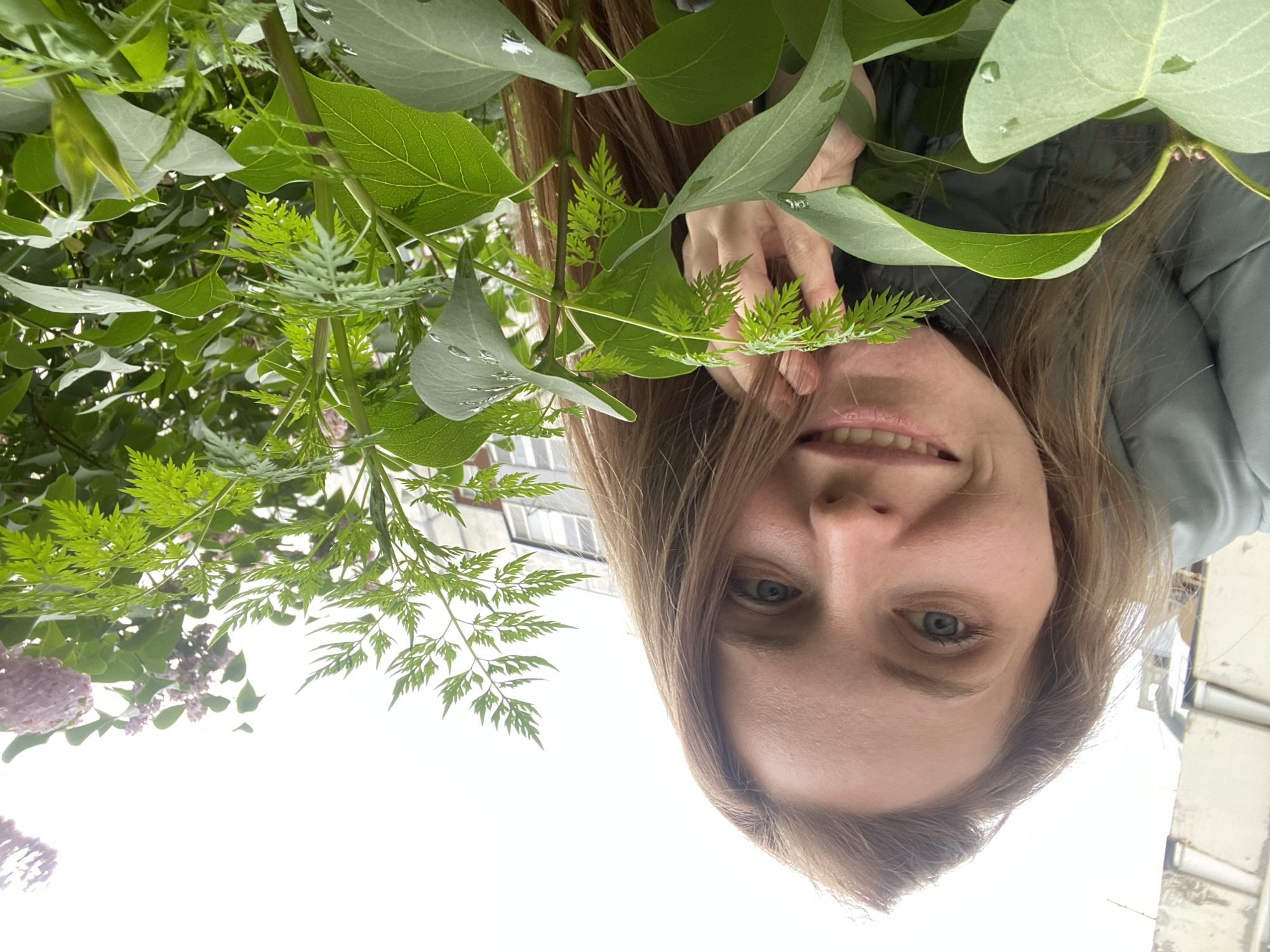 A medium shot shows a woman with long brown hair and blue eyes, looking directly at the camera with a slight smile. She is wearing a light green puffer jacket. Her right hand is touching her face near her mouth, with her fingers partially obscuring a feathery green leaf. The background consists of lush green foliage, including some larger leaves and smaller, fern-like leaves, and hints of pink blossoms in the upper right corner. In the distance, there are buildings, suggesting an urban environment. The lighting is overcast, indicating a cloudy day. The scene appears to be outdoors, possibly in a park or garden area in Yuzhno-Sakhalinsk, Russia, given the architectural style of the distant buildings which resembles those found in Russian cities.