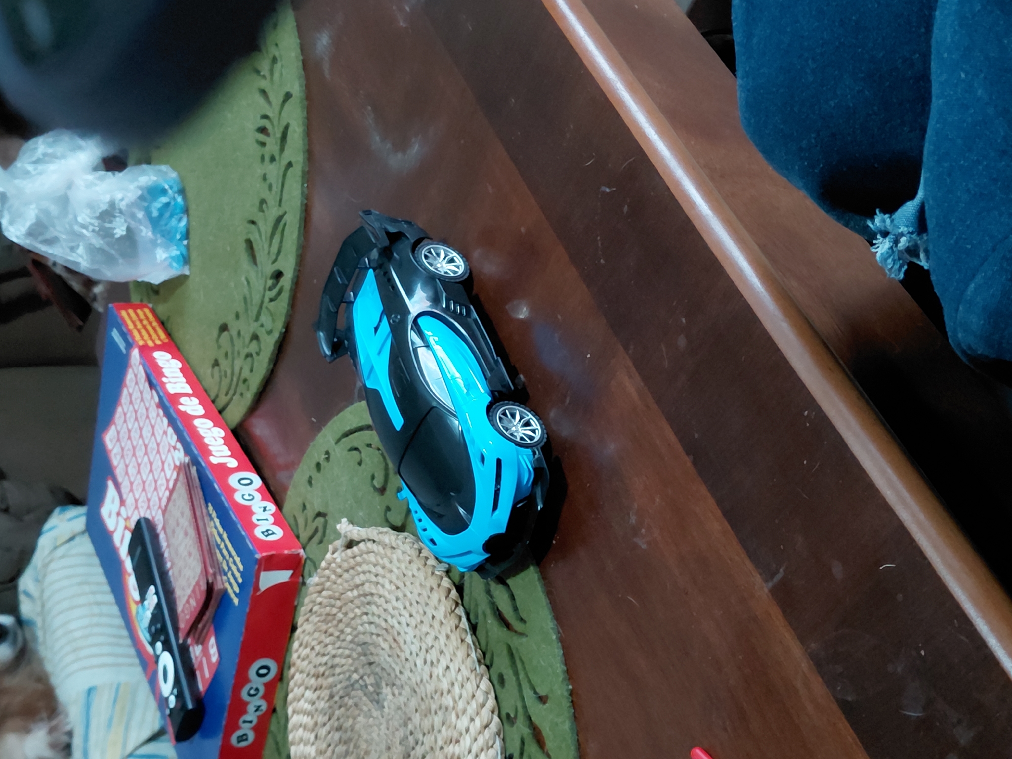 The image depicts a scene on a brown wooden table. A blue and black toy car rests on the table. Next to the car is a Bingo board game box and a round woven mat. A plastic bag is on the left of the bingo game. A blue upholstered chair is on the right side of the frame. The location in the city of Santa Ana, Venezuela, is unknown.