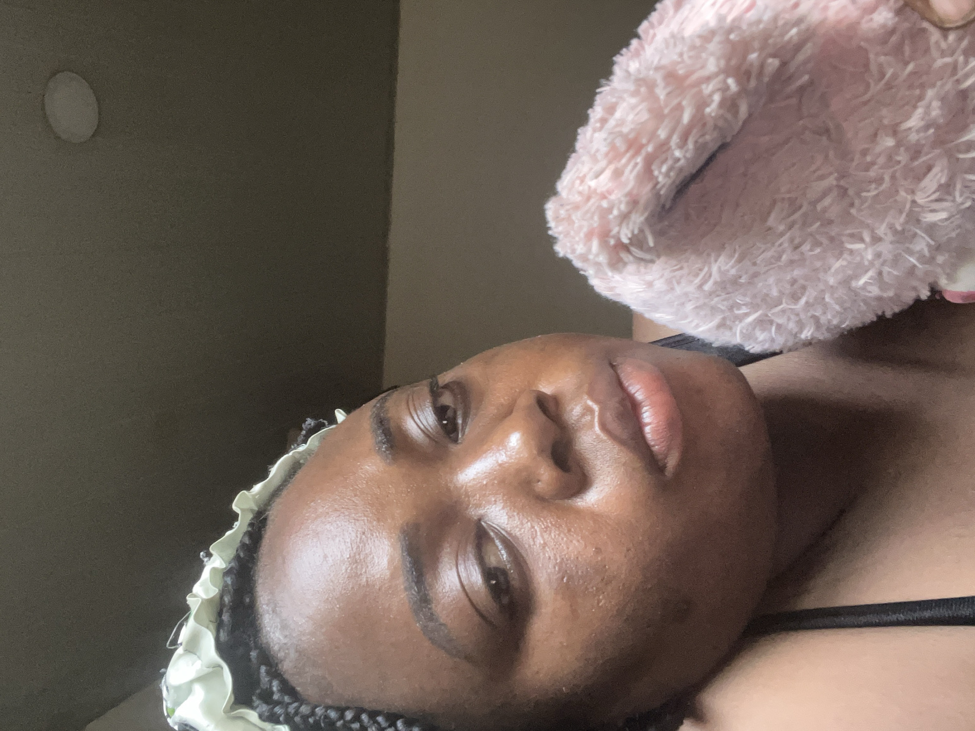 The image presents a close-up portrait of a person, likely a woman, with dark skin and dark braided hair, appearing to be lying down or reclining. Her face is directed towards the camera, showcasing defined eyebrows, dark eyes, and full, slightly parted lips with a natural sheen. Her skin appears smooth and moisturized.
Around her head, a light-colored, ruffled fabric, resembling a bonnet or head covering, is visible. In the upper right of the frame, a fluffy, soft pink object, made of a plush material, is positioned near her head or shoulder. A black strap, consistent with a tank top or bra, is visible across her chest in the lower right.
The background consists of a plain, muted wall in shades of grey or beige. A circular, light-colored object, possibly a light fixture or a decorative element, is visible on the upper left portion of the wall. The lighting is soft and ambient, indicating an indoor setting. The individual's expression is neutral and relaxed. No text is visible in the image. The location context is Abuja, Nigeria.
