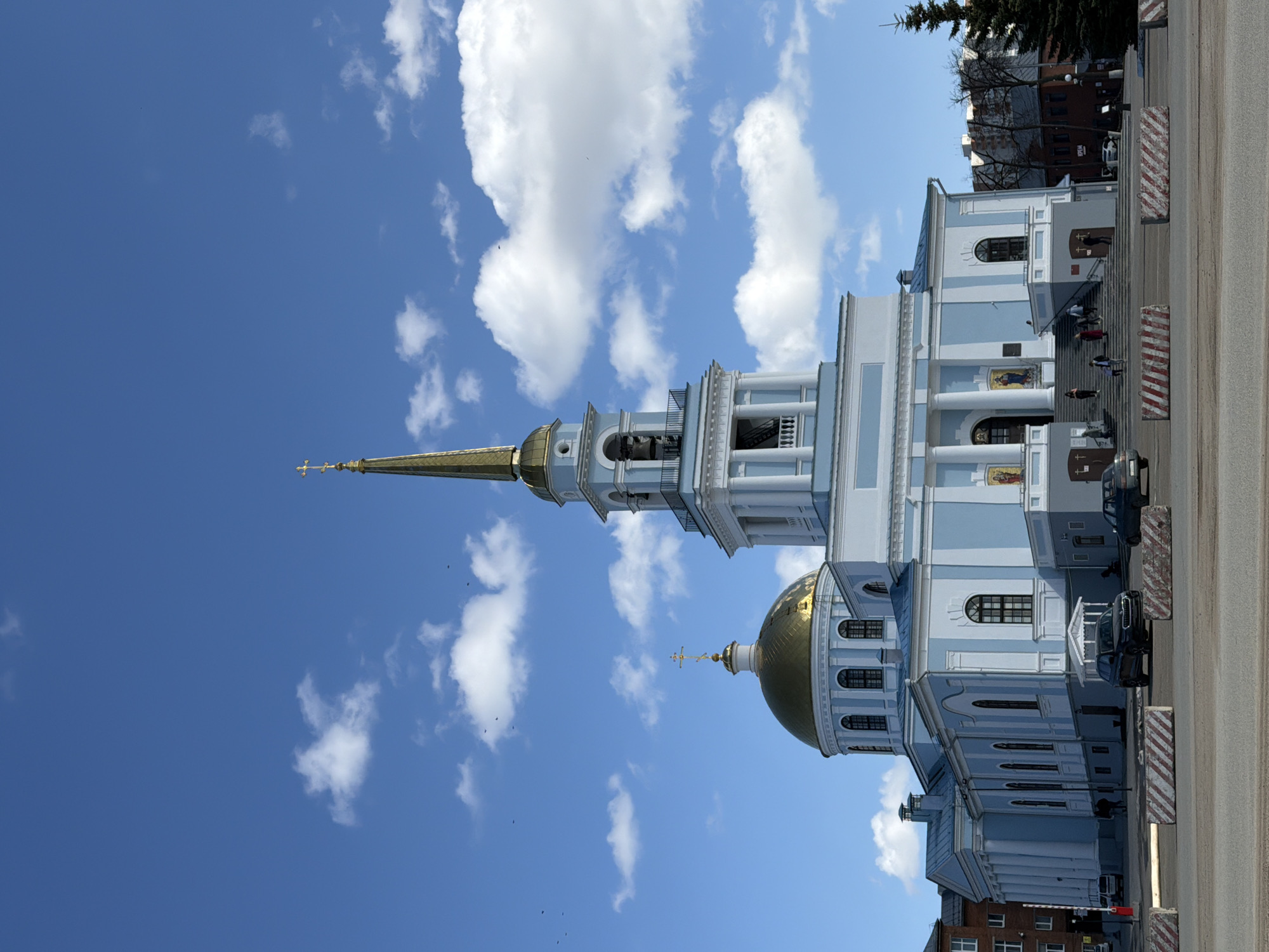 The image depicts the Annunciation Cathedral in Votkinsk, Russia. The church is a light blue and white structure with a gold dome and spire. The sky is blue with white clouds. There are a few people visible on the steps of the cathedral, and several cars parked nearby. The cathedral is located on a street in Votkinsk.