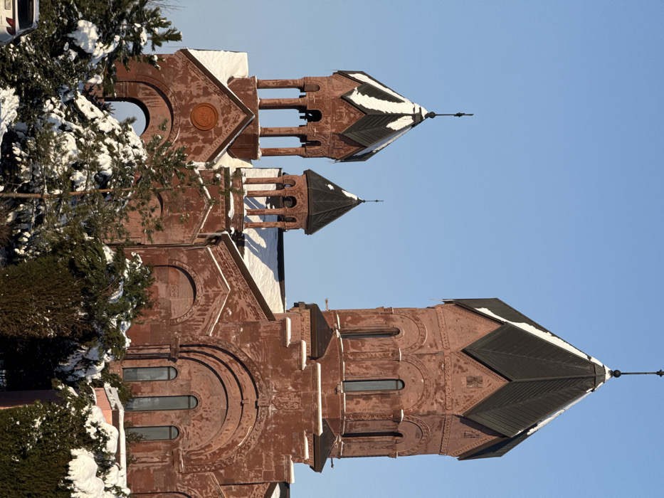 The image depicts the Surp Hovhannes Mkrtich (Saint John the Baptist) Armenian Apostolic Church in Rassvet, Russia, under a clear, bright blue sky during winter. The prominent structure is constructed from reddish-brown brick or stone, displaying intricate architectural details.
The church features a large central conical dome with a dark, segmented roof, topped by a slender cross finial. This main dome section includes tall, narrow arched windows. To the left of the central dome, two smaller, open bell towers with dark conical roofs are visible, each also topped with a cross finial. These bell towers feature arched openings supported by columns. An additional smaller, closed dome is partially visible behind these bell towers. All visible roofs and various architectural ledges on the building are partially covered with white snow. Decorative carvings, including cruciform designs, are discernible on the church's facade and dome bases.
In the foreground, a dense collection of evergreen trees and bushes, heavily covered with white snow, partially obscures the lower sections of the church. A portion of a white vehicle, possibly an SUV, with a partial license plate ending in "...782," is visible in the bottom-left corner, largely obscured by the snow-laden foliage. The scene is illuminated by strong, directional sunlight, indicating a bright winter day. No individuals or active interactions are present in the frame.