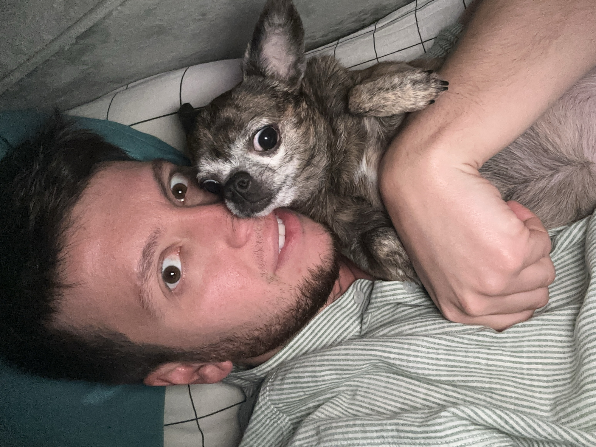 In this close-up, overhead shot, a man and a small dog are lying down together. The man, with short dark hair and a beard, is looking directly at the camera with wide eyes. He is wearing a light green and white striped button-down shirt. The dog, a small breed with brown and grey fur and large eyes, is nestled against the man's chest and shoulder, also looking towards the camera. They are in a horizontal position, possibly in bed, as suggested by the pillow with a black and white grid pattern visible in the background. The lighting is dim, creating a soft ambiance. No specific location in Mirny, Russia, can be determined from the image.