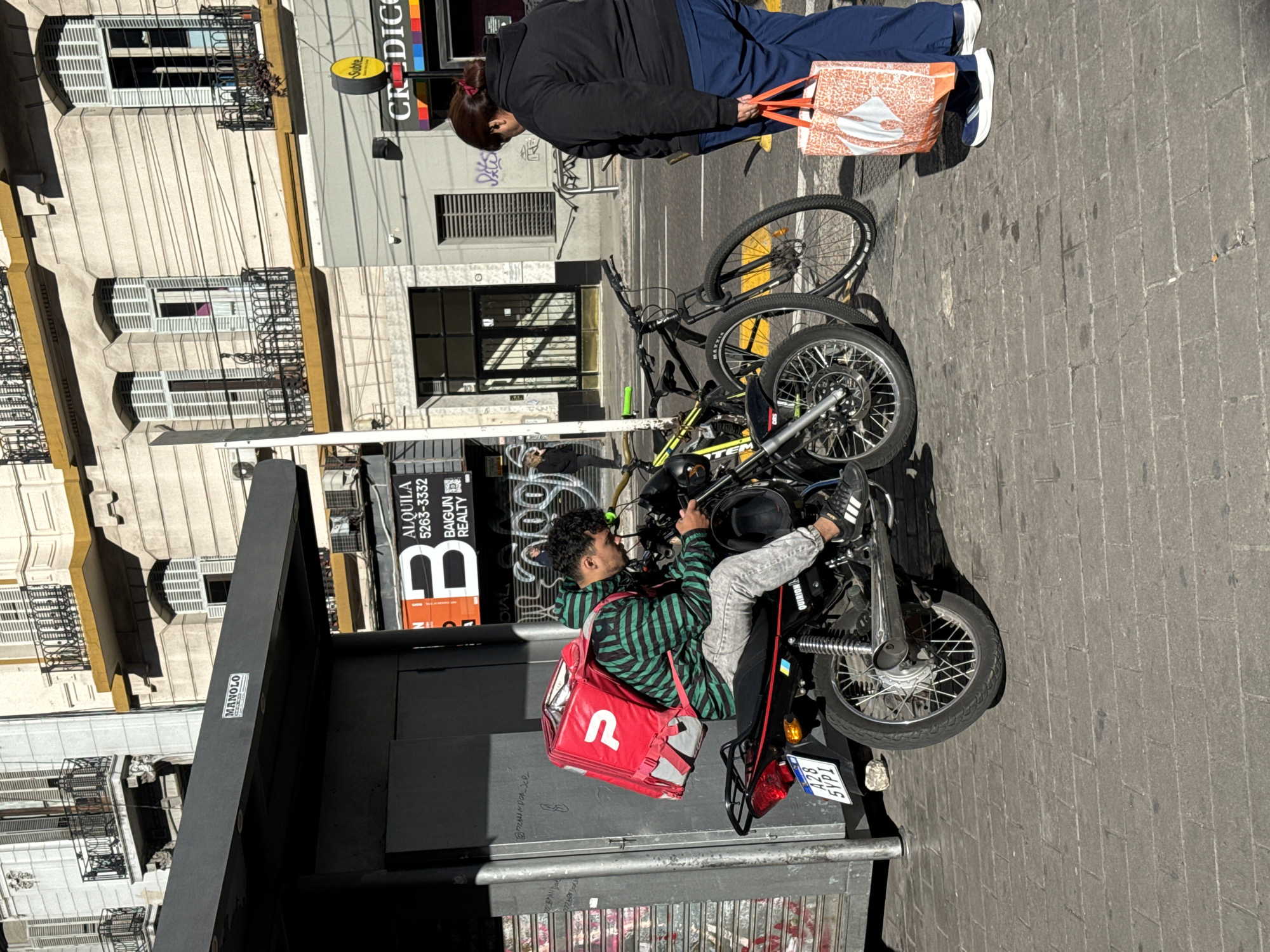 The image depicts a street scene in Buenos Aires, Argentina. A delivery person sits on a small motorcycle, wearing a red delivery backpack. They are parked next to a bus stop, with posters visible. Behind them is a black bicycle and an older woman with a shopping bag walking perpendicular to the delivery person. The buildings in the background are multistory and appear older, with arched windows and decorative balconies. The street is paved with brick.
The license plate of the motorcycle shows A28 SYPI. The bus stop has a sticker with the name MANOLO.
A sign behind the bus stop reads "ALQUILA 5263-3332, BAIGUN REALTY."
A sign on a building reads "Subte CRIDICO."