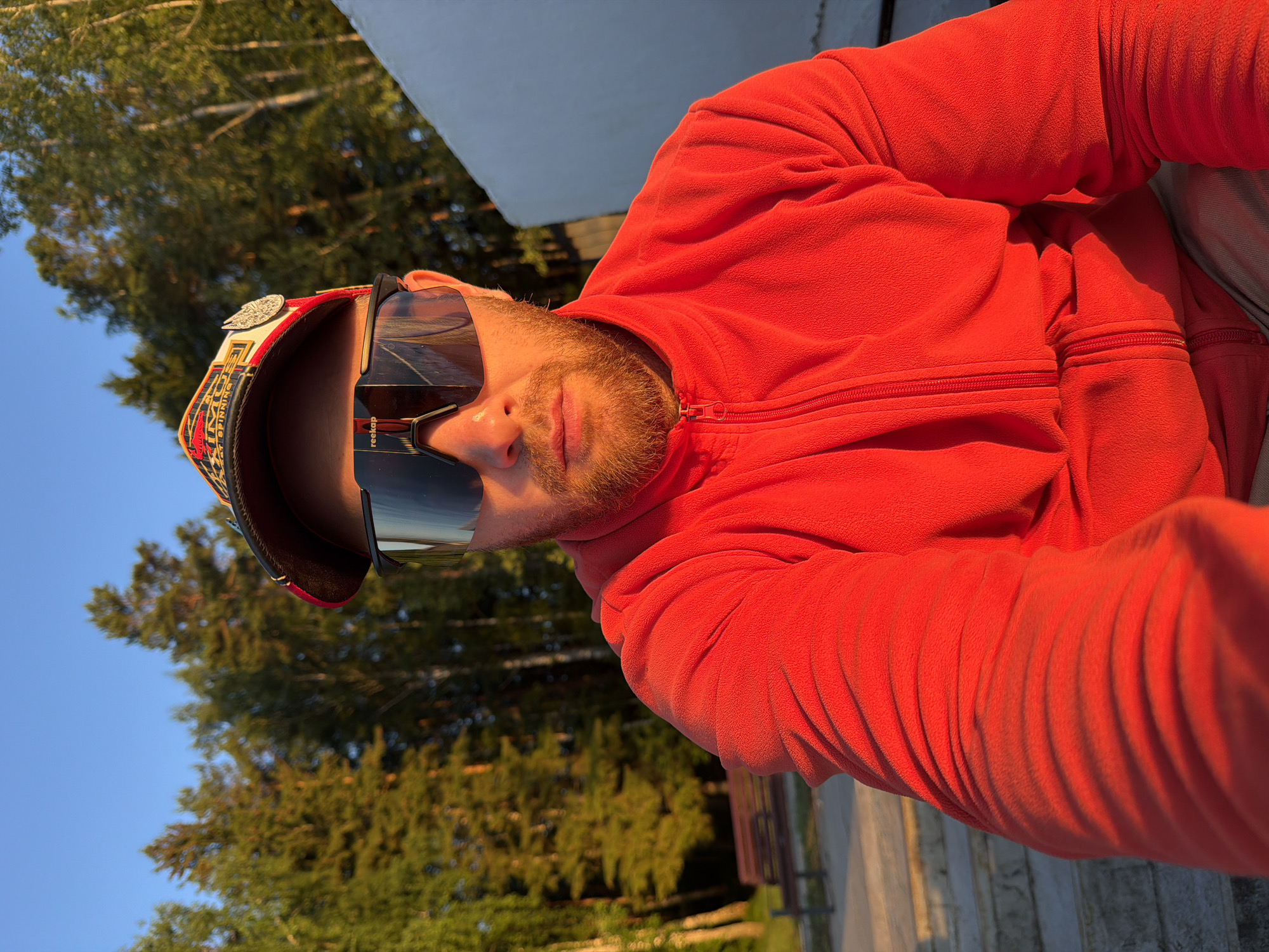 A man with a beard and reddish-brown hair is captured in a selfie from the chest up, facing the camera directly. He is wearing a red zip-up fleece jacket and a baseball cap with a colorful pattern and a circular emblem on the front. His eyes are obscured by large, dark, wraparound sunglasses with a reflective visor. The background consists of a stand of tall green trees, bathed in warm, golden sunlight, suggesting it is either sunrise or sunset. To the right of the man, a large, rough-textured white surface is visible, possibly a wall or a building. In the lower left of the frame, a portion of concrete stairs and a wooden bench are discernible, indicating an outdoor public space. The setting appears to be a park or recreational area, possibly within or near the city of Chaikovsky, Russia, given the presence of birch and pine trees typical of the region.