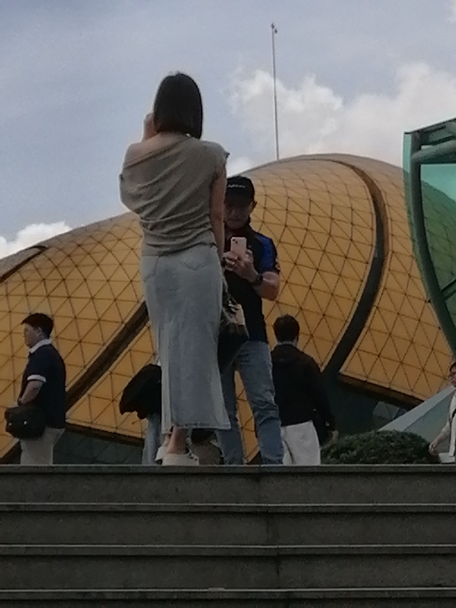 The image captures a group of people on a set of concrete stairs outside a building with a distinctive golden, geometric dome. A woman in a light gray off-the-shoulder top and a long, light blue denim skirt stands in the foreground, facing away from the camera and holding a phone to her ear. To her right, a man wearing a black baseball cap, a dark blue and black shirt, and blue jeans holds a pink smartphone, appearing to take a photo. Behind them, another person in a black jacket and white pants stands near a lush green bush. To the left, a person in a dark blue polo shirt and dark pants, carrying a dark bag, looks to the side. In the background, the golden dome building, which resembles the exterior of the Da Lat Flower Garden's Exhibition Center, is partially visible against a cloudy sky. The scene suggests a public outdoor space, likely a tourist attraction in Da Lat, Vietnam, with people engaged in activities like taking photos and socializing.