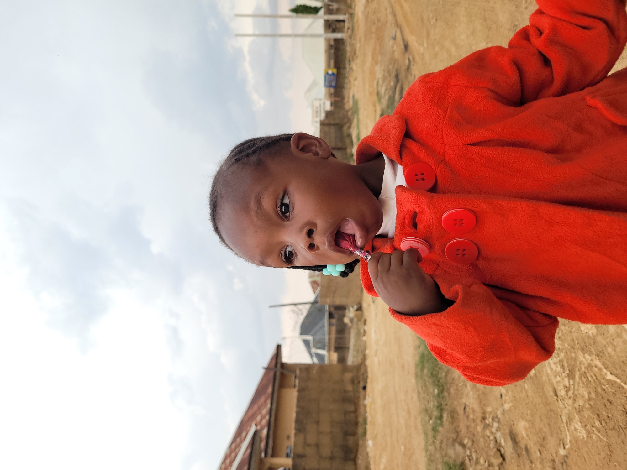 A young child stands in an outdoor scene near Barakin Akawo, Nigeria. The child wears a bright orange coat with large red buttons. The child is holding a lollipop in hand and partially in the mouth. Buildings can be seen in the background, with dirt and grass visible in the foreground. The sky is overcast.