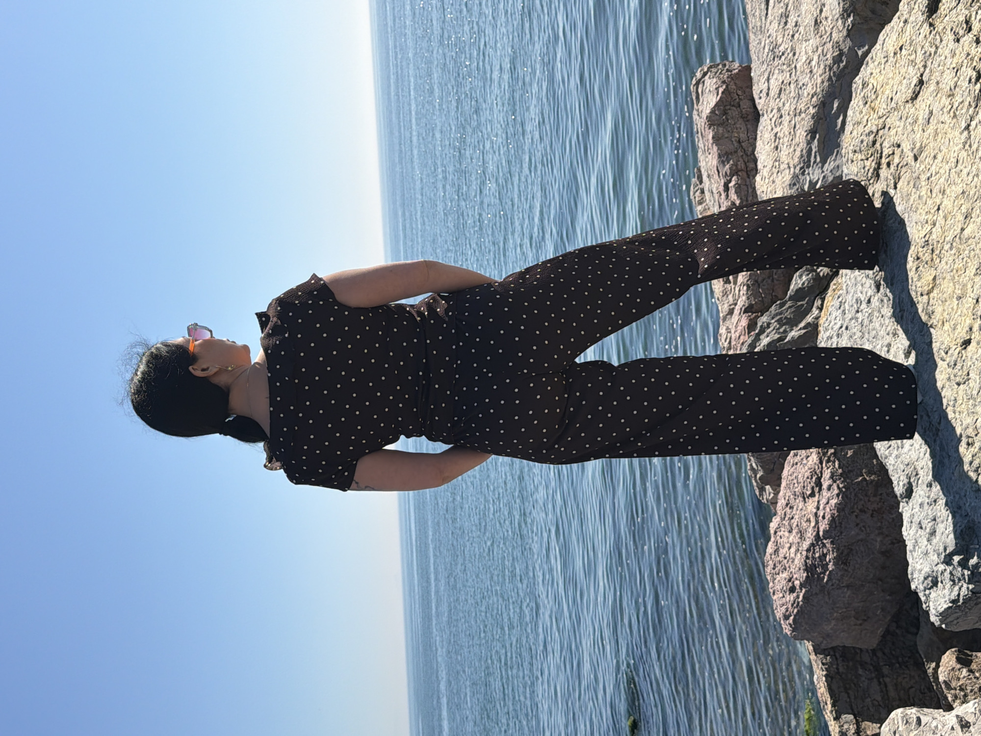 A full shot shows a person standing on a rocky shoreline with the sea and sky visible. The person, a woman, is seen from behind. She is wearing a brown jumpsuit with white polka dots and sunglasses. She stands with her hands on her hips. The sea behind her is calm and blue, with the sky above it clear and light blue. The rocks on the shoreline are grey and tan. The location is likely in Kadıköy, Turkey, given the landscape.