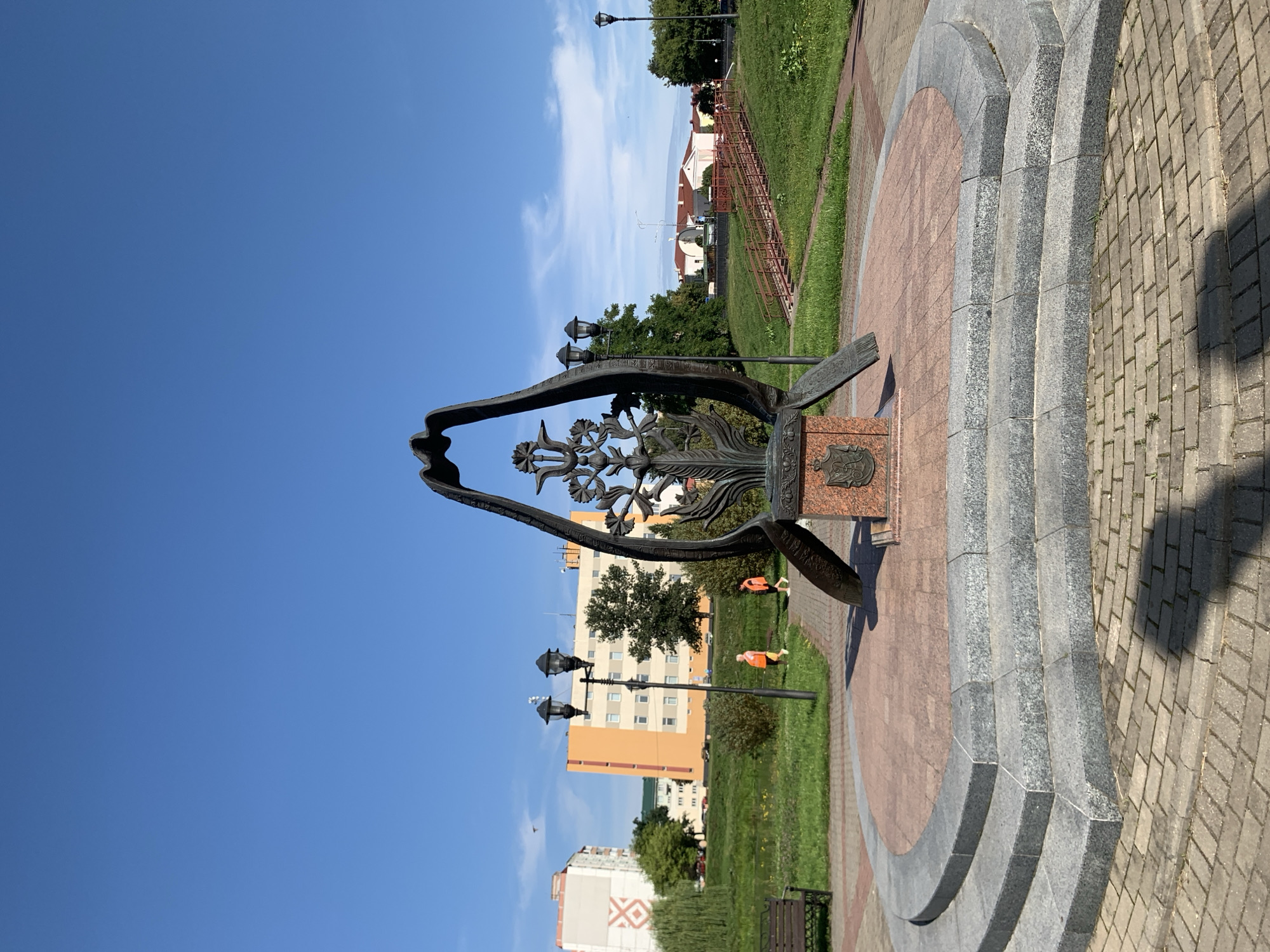 The image captures a monument in Slutsk, Belarus, with a clear blue sky overhead. The monument, made of dark metal, features an intricate, tree-like structure enclosed within curved, wing-like elements. It stands on a tiered, circular base constructed of stone and patterned bricks. The monument itself is mounted on a granite pedestal with a crest. In the background, there are buildings, green spaces, and a pathway with stairs. Two individuals wearing orange vests are walking on the pathway in the middle ground, and a park bench is visible to the left. The lighting suggests it is daytime.