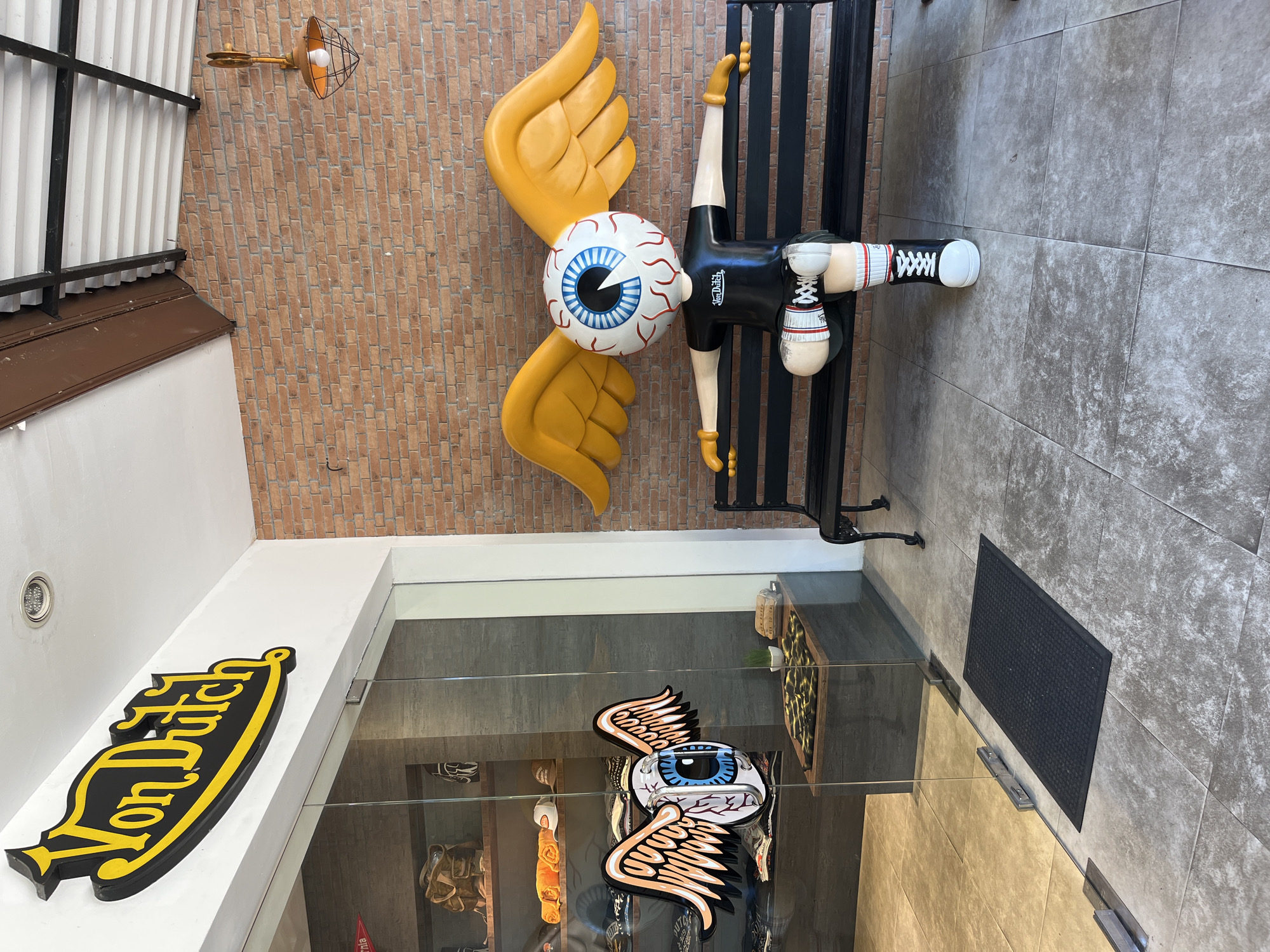 The image depicts the exterior of a Von Dutch store in Kuta Selatan, Indonesia. A prominent yellow and black Von Dutch logo is mounted above the glass entrance. To the right of the entrance, a large, quirky statue of a winged eyeball character wearing a black t-shirt, shorts, striped socks, and high-top sneakers sits on a black bench against a brick wall. The statue has its arms outstretched. The store entrance features glass doors with a decal of a winged eyeball on the handle. Inside the store, shelves display merchandise, including bags and apparel. The ground is tiled with large grey square tiles, and a black doormat is placed at the entrance. The environment appears to be outdoors, with natural light illuminating the scene.